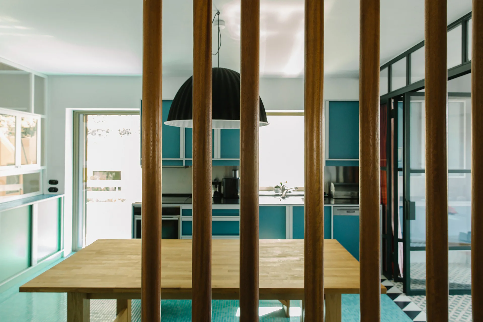 A modern kitchen with blue cabinets and a light wood dining table, seen behind a partition of vertical wood slats.