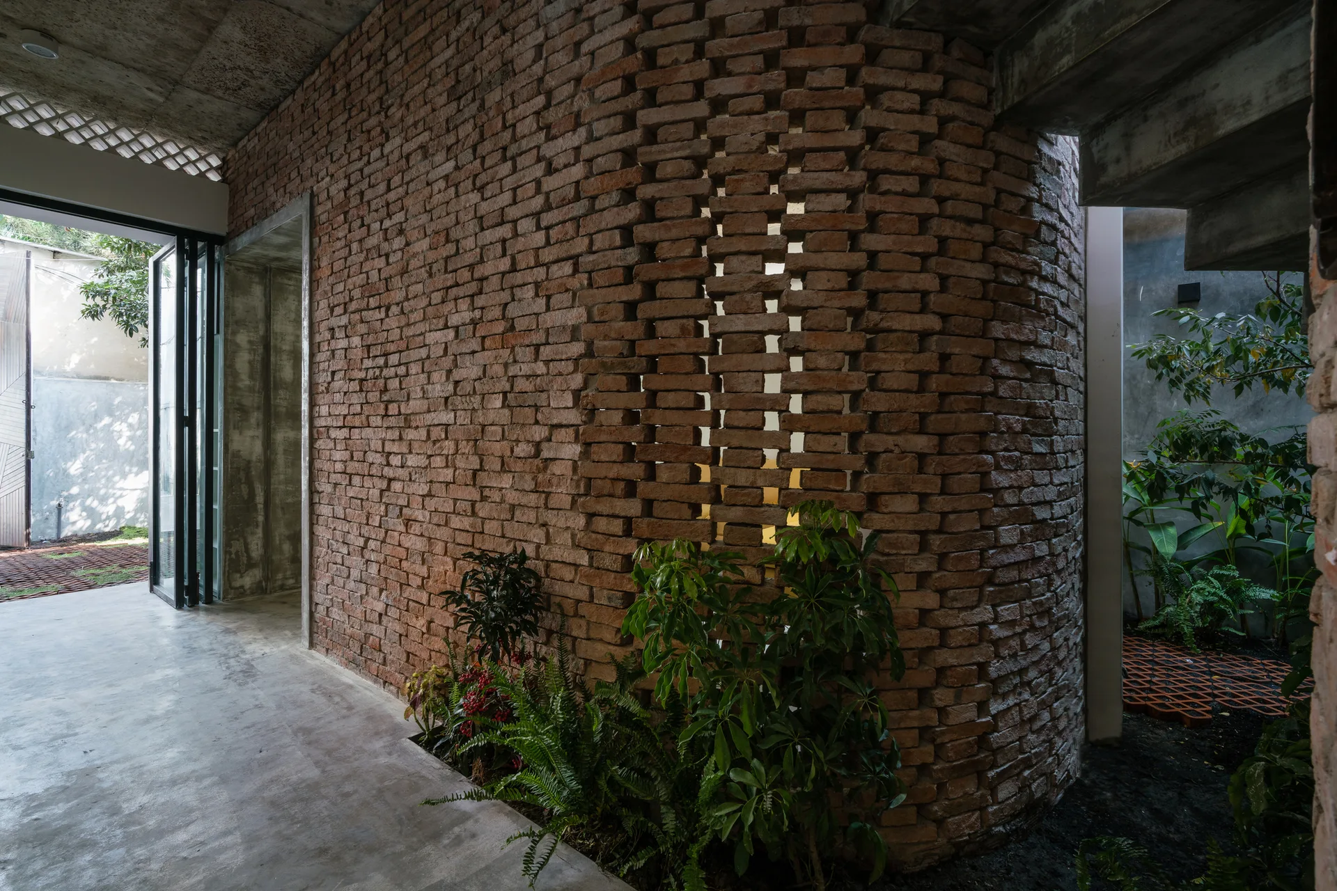 A curved wall of exposed red brick with open perforations sits next to a raw concrete floor and ceiling, accented by green plants.