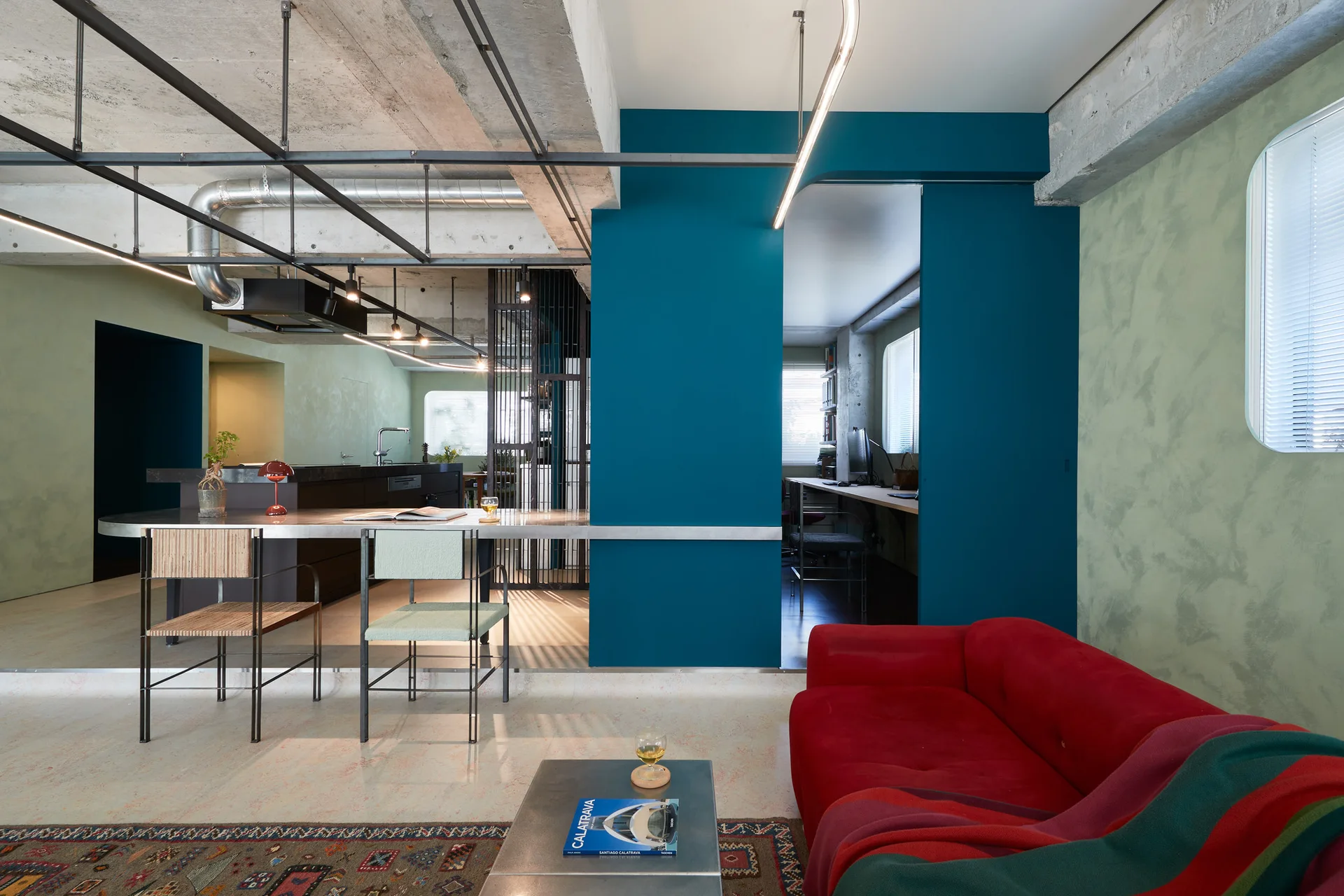 A modern open-plan living room features a bright red sofa, a metal-topped kitchen island, a deep teal partition wall, and an exposed concrete ceiling.