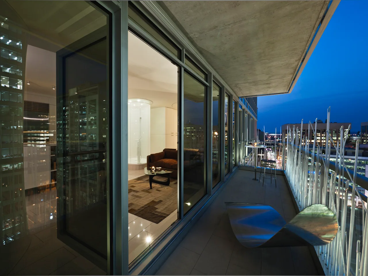A night view of a modern high-rise apartment balcony featuring a curved metal lounge chair, sliding glass doors, and a decorative rod railing.