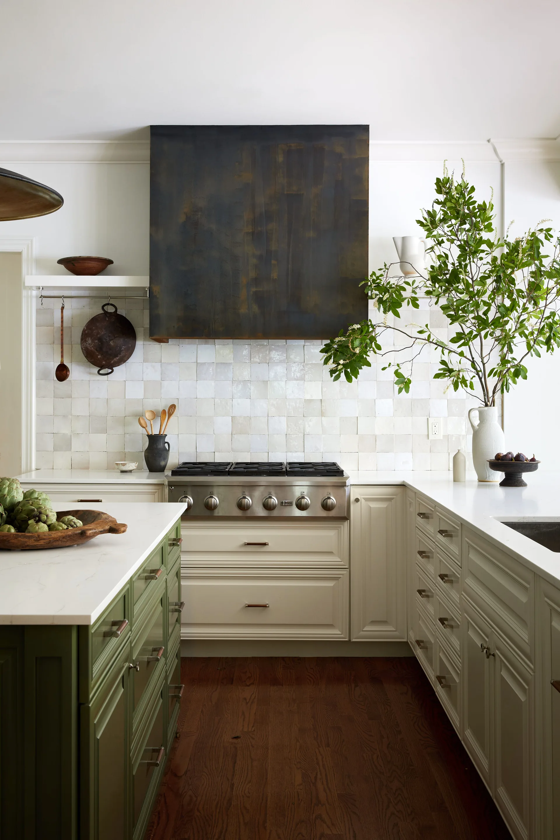 A kitchen features a dark metal range hood over a stainless steel stove, glossy white tile backsplash, cream cabinets, and a sage green island.