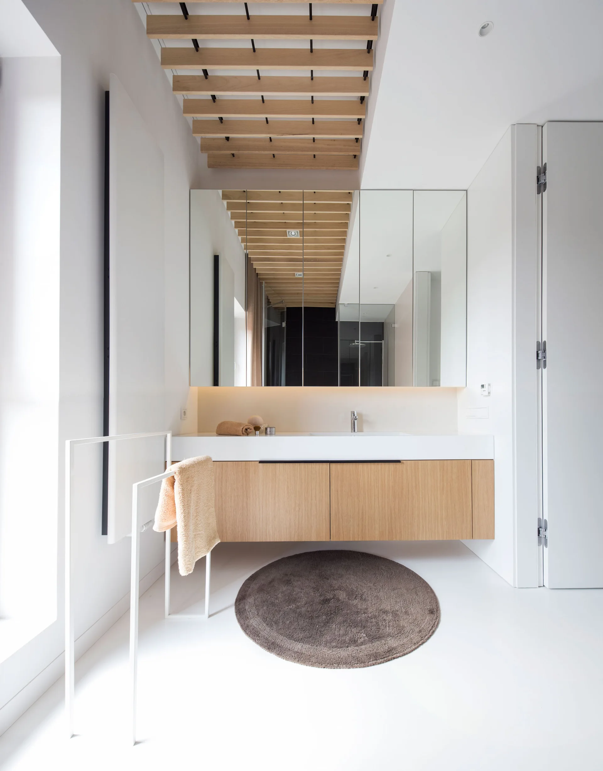 A modern white bathroom with a floating light wood vanity, a large mirrored medicine cabinet, a circular brown rug, and wooden ceiling slats.