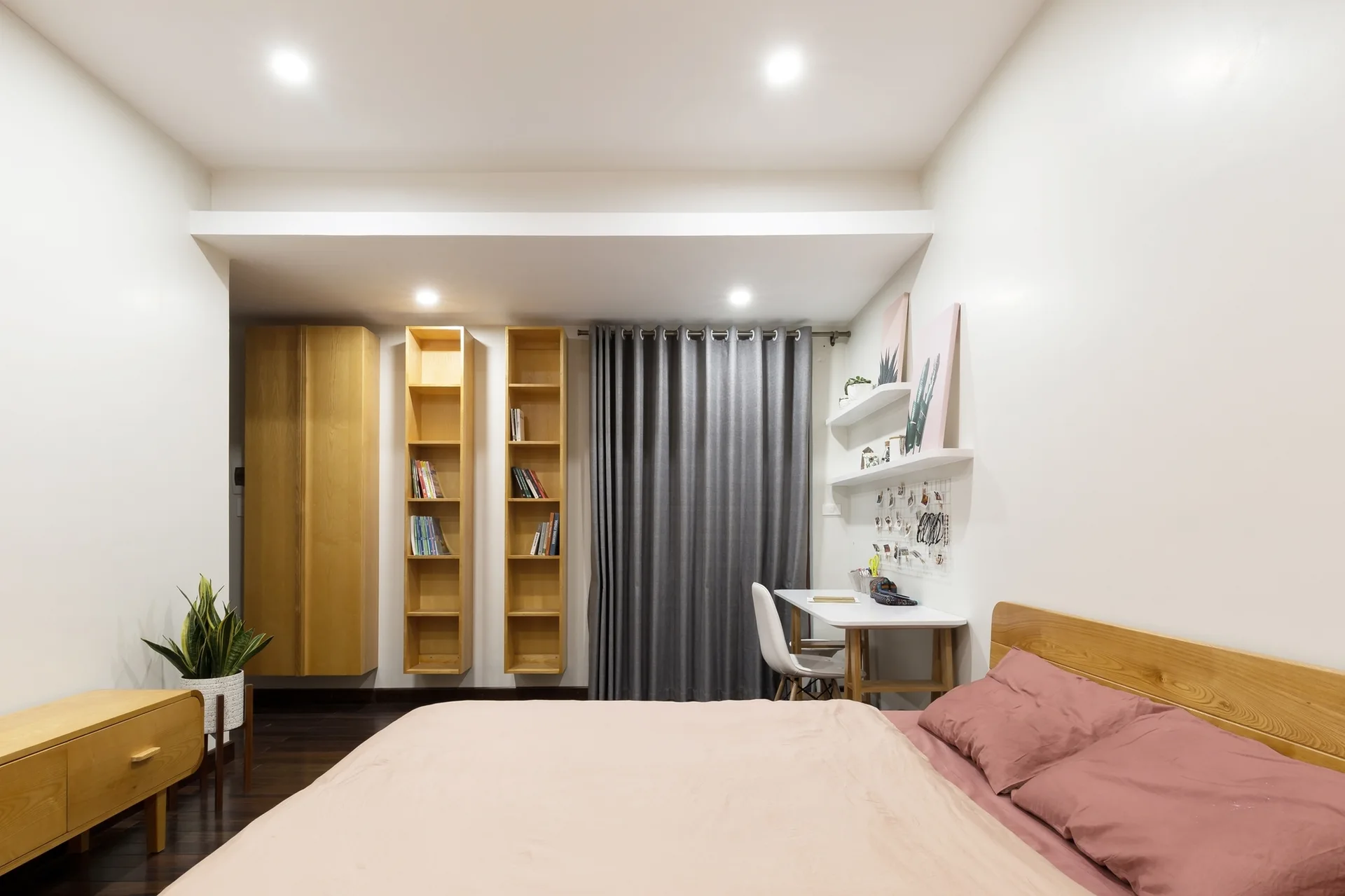 A minimalist bedroom features a wooden bed with blush pink pillows, a white study desk with a chair, and vertical wood bookshelves mounted to the wall.