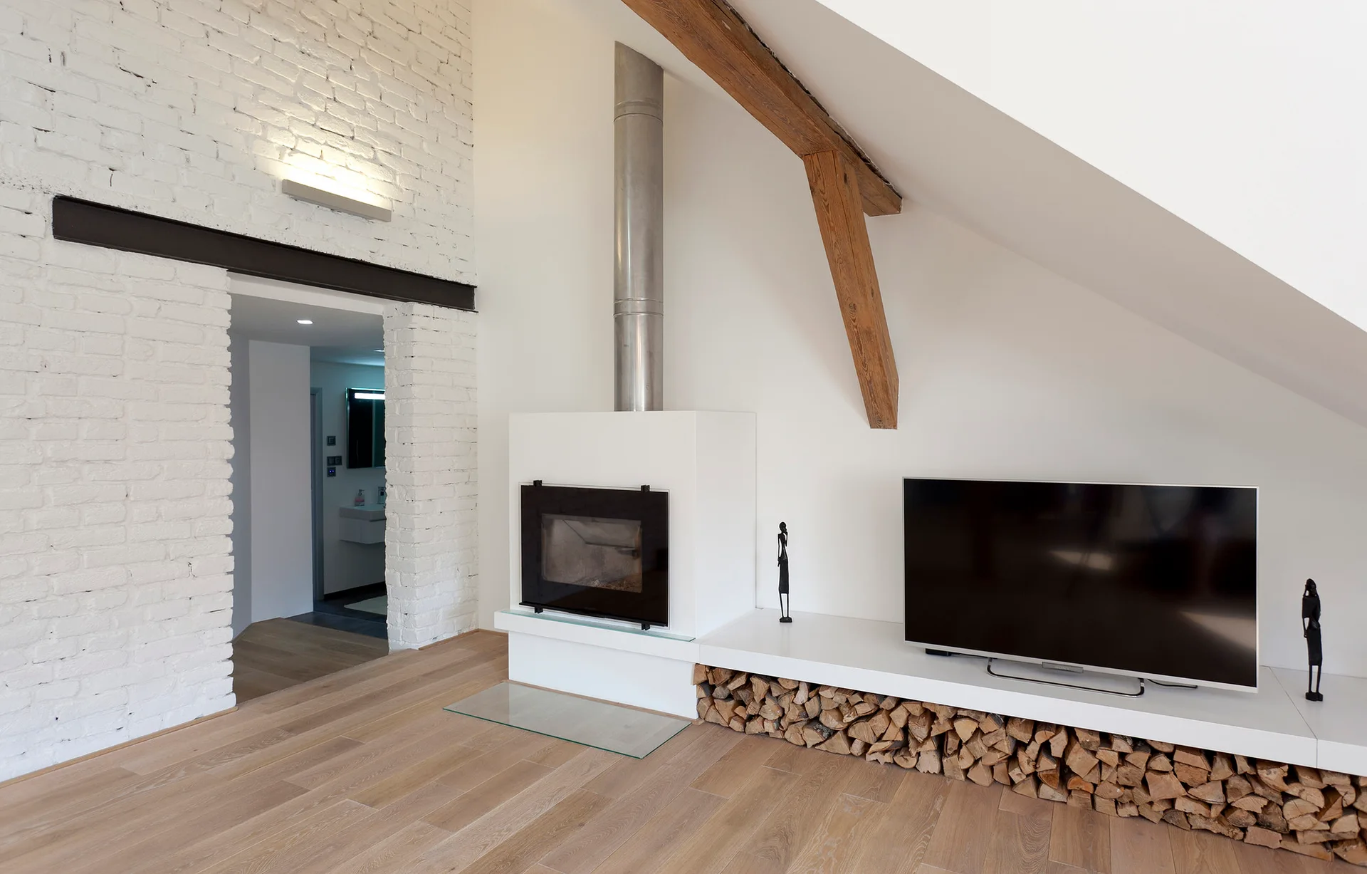 A modern attic living room featuring a white brick wall, a low built-in fireplace ledge with stacked firewood beneath, and a sloped ceiling.