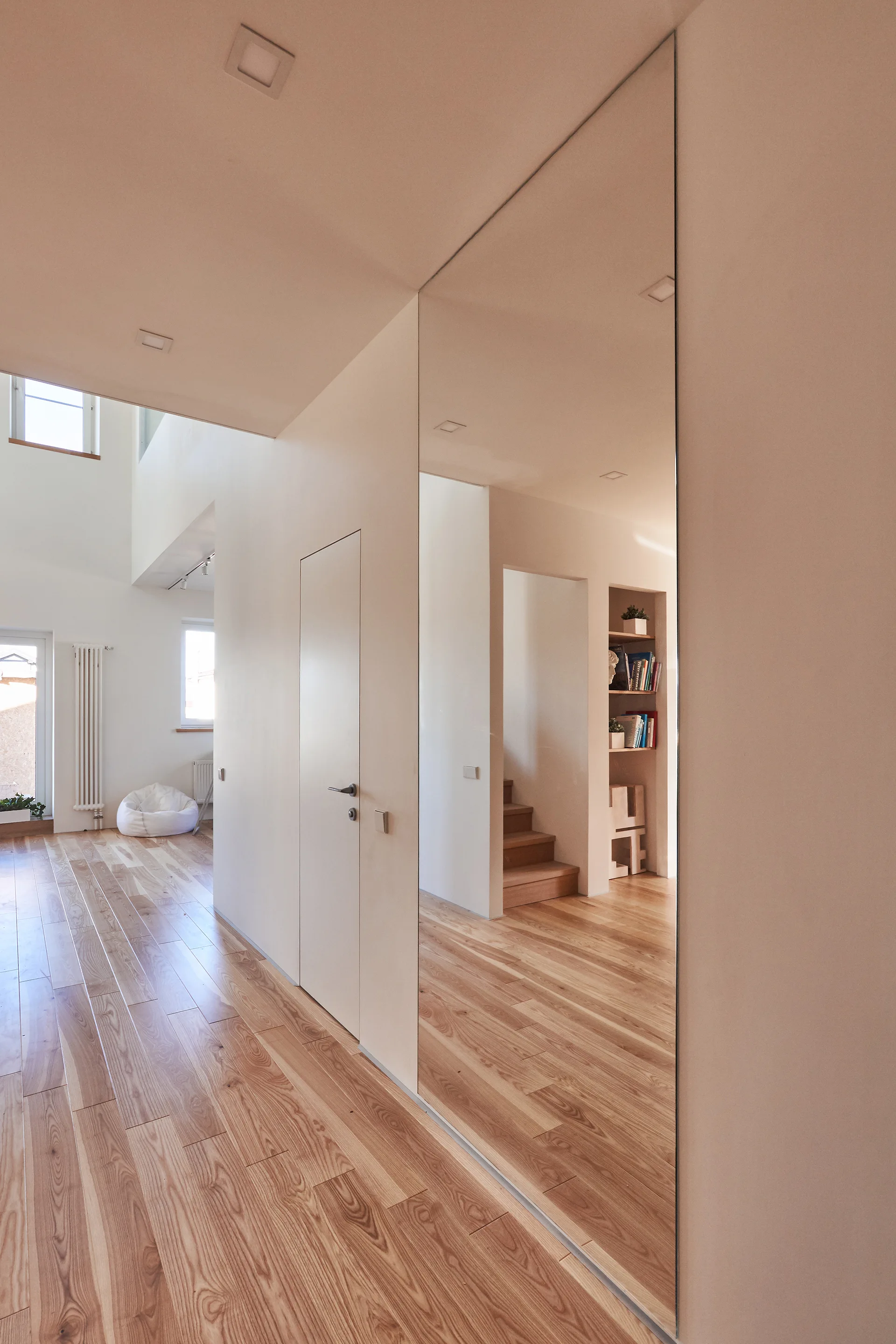 A minimalist hallway with light wood flooring features a flush door and a large frameless floor-to-ceiling mirror reflecting a wooden staircase.