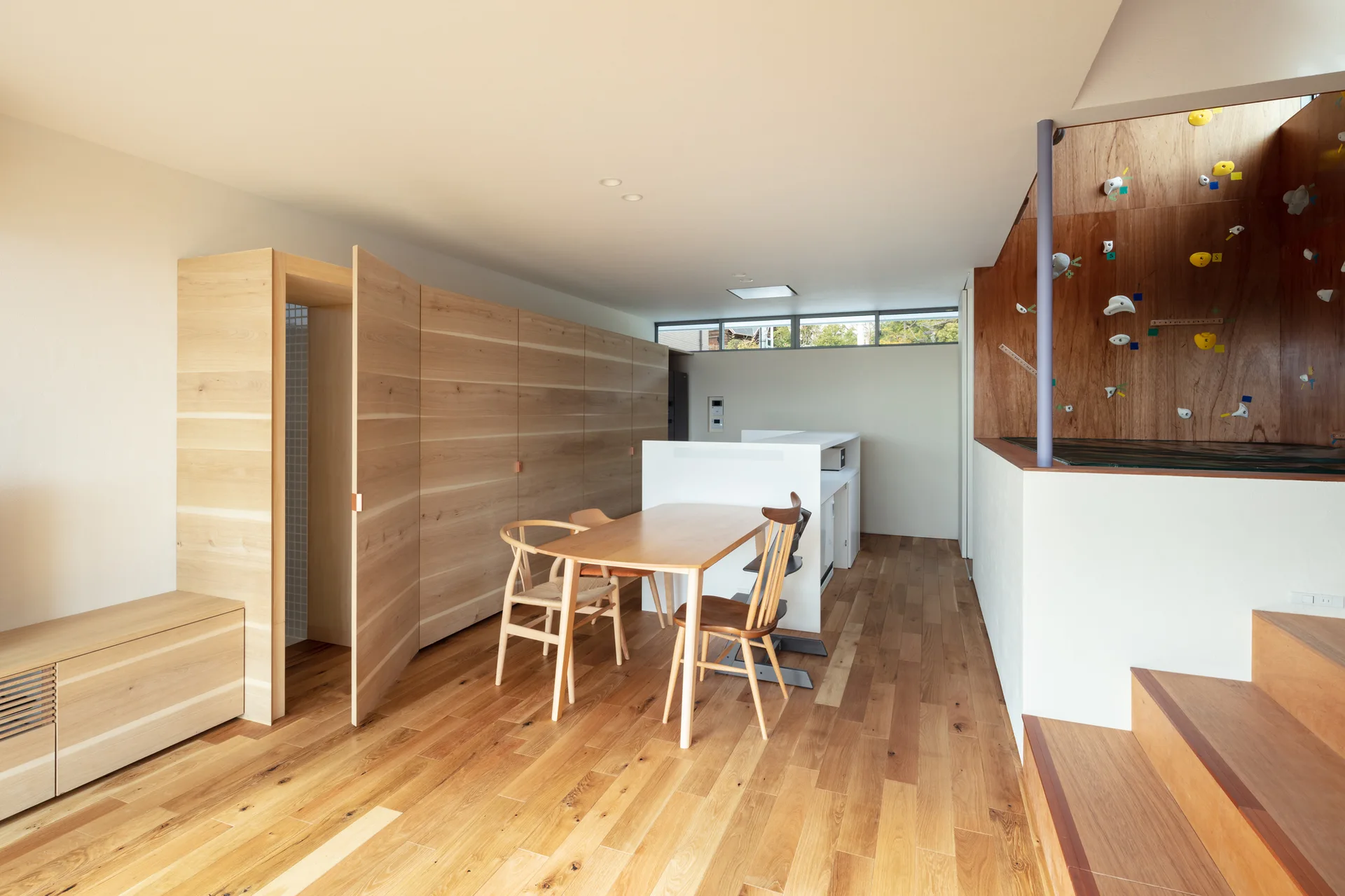 A minimalist light wood dining room with matching table and chairs, seamless built-in storage cabinets, and an indoor climbing wall next to the stairs.