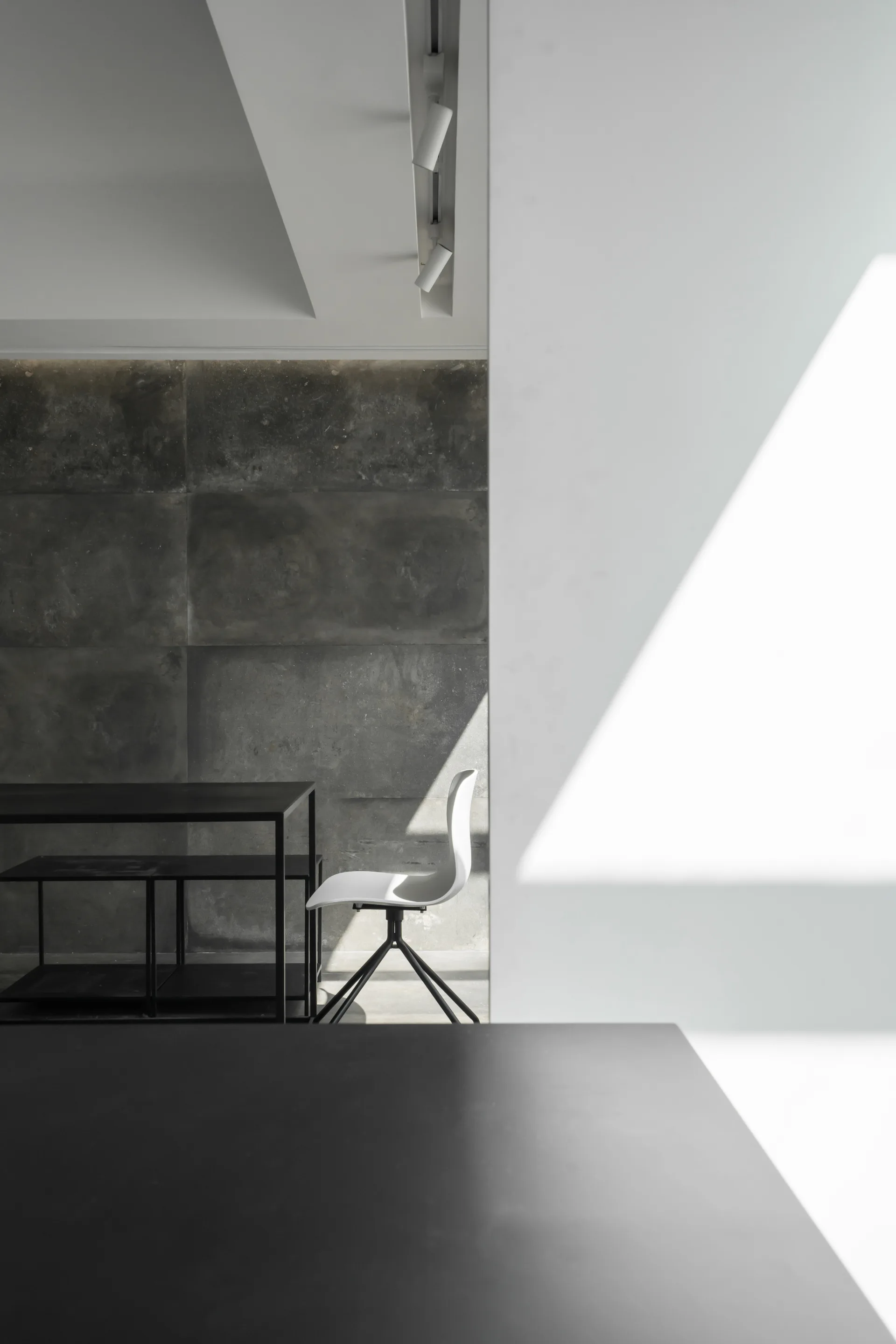 A white desk chair and black table set against a dark grey textured paneled wall and a smooth white partition.