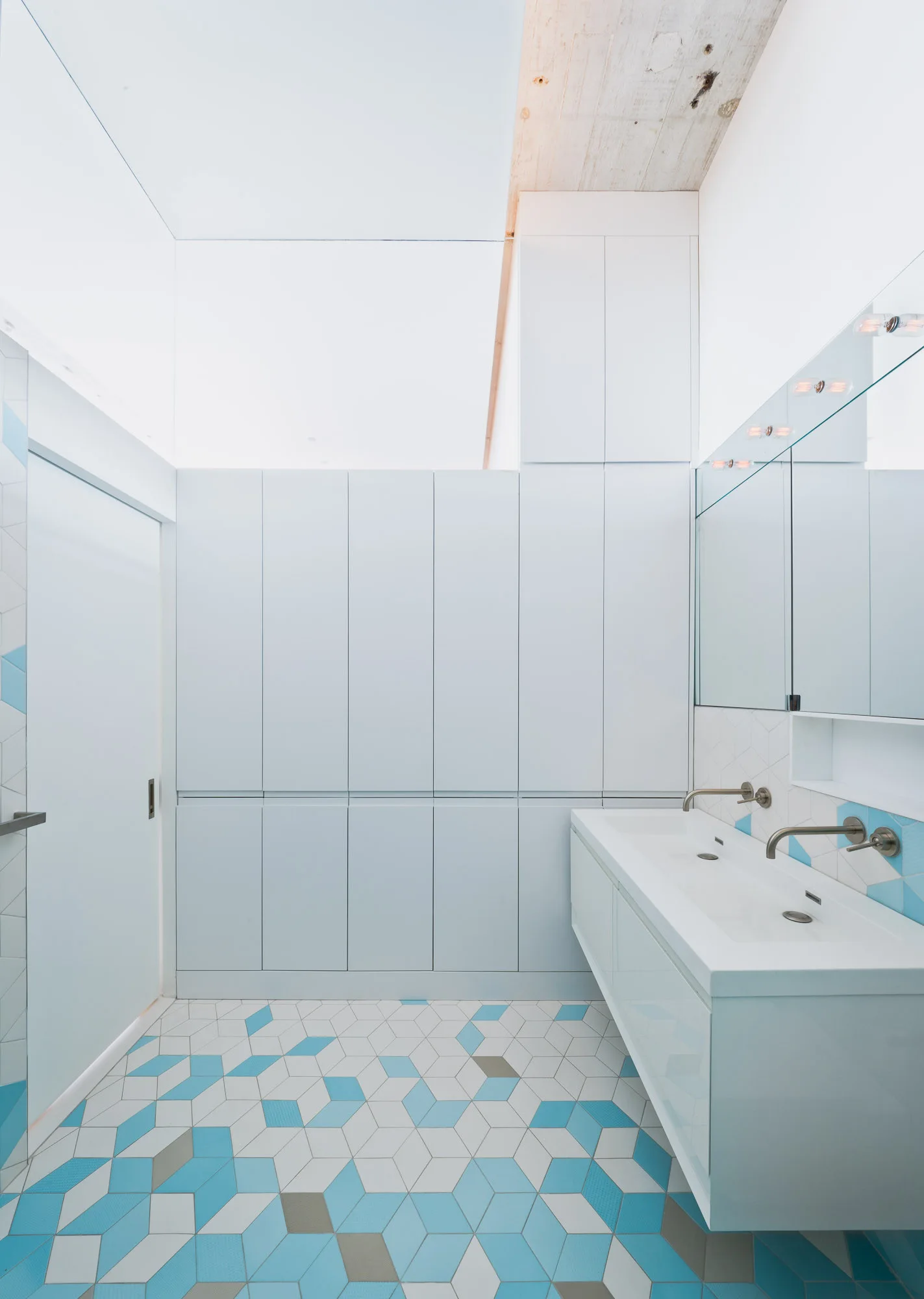 A modern bathroom features a long white wall-mounted vanity, pale blue cabinetry, and a geometric blue and white patterned tile floor.
