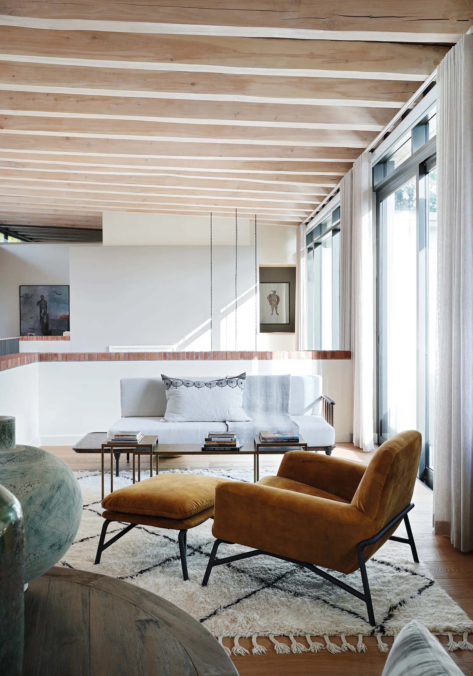 A bright living room features an exposed wood beam ceiling, a mustard yellow velvet armchair with ottoman, and a grey sofa on a diamond pattern shag rug.