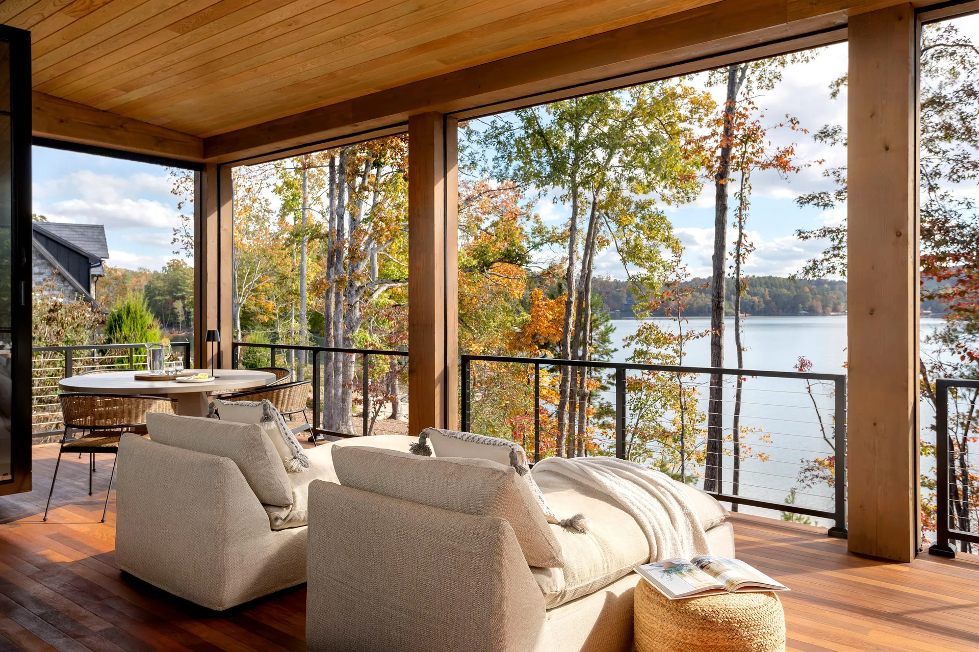 A covered wooden porch features beige upholstered lounge chairs, a black cable railing, and a view of a lake bordered by autumn trees.