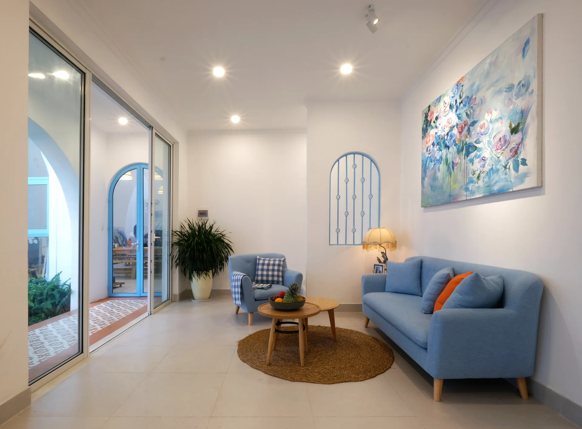 A bright living room features a blue sofa and armchair, a round jute rug with wooden coffee tables, and a large floral painting on white walls.