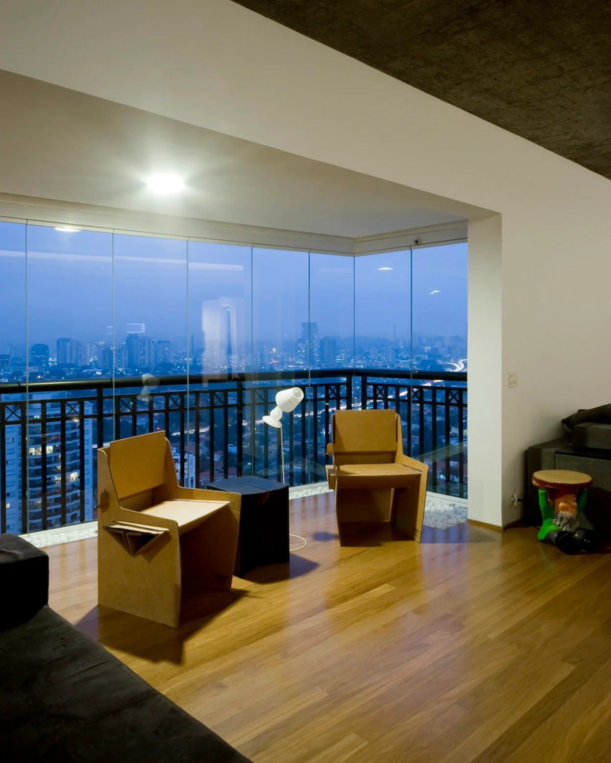 Two geometric cardboard chairs and a small black side table sit on wood floors next to a panoramic glass wall overlooking a city at dusk.