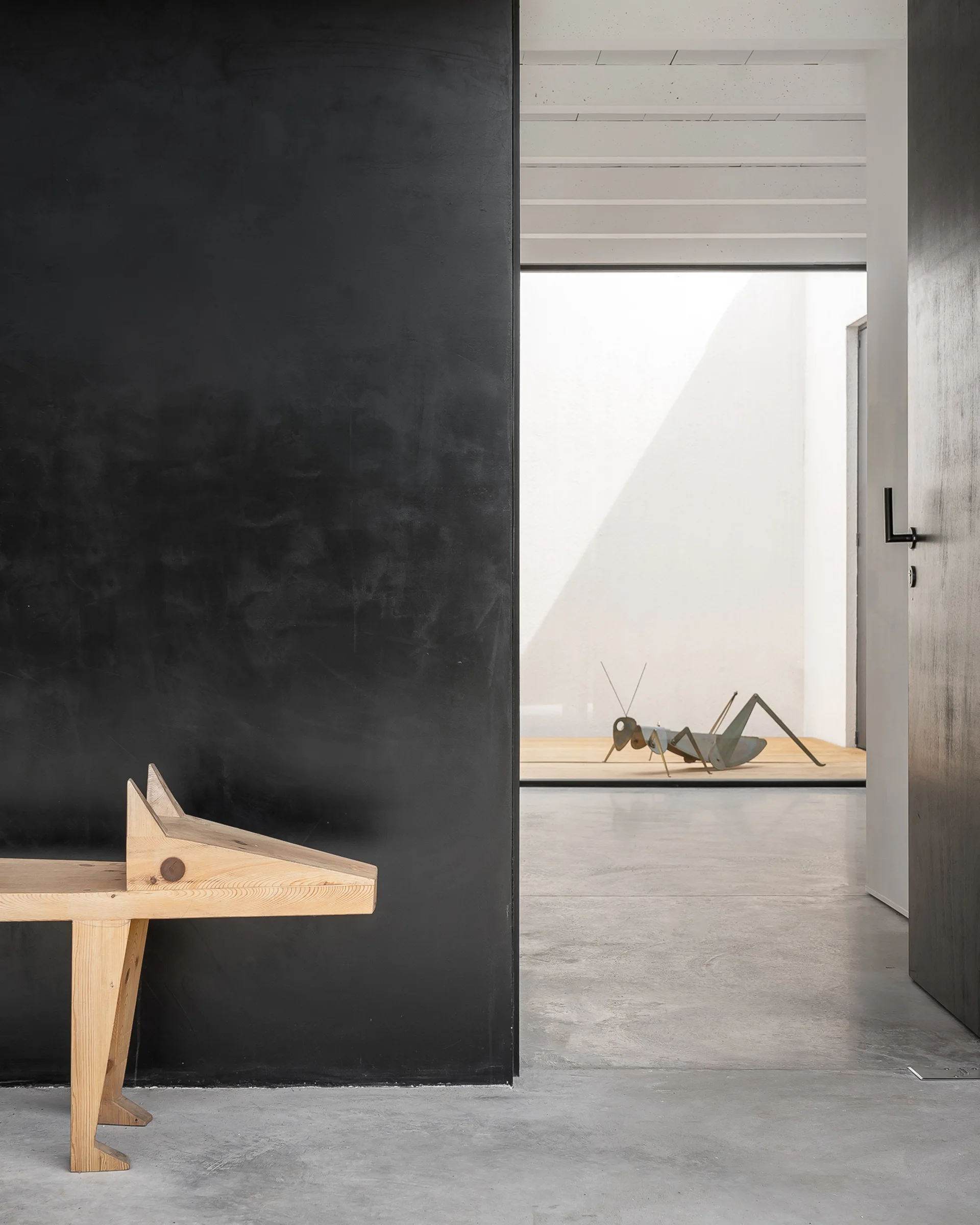 A minimalist entryway features a light wood animal sculpture next to a matte black wall, looking out toward a bright courtyard with a metal grasshopper.