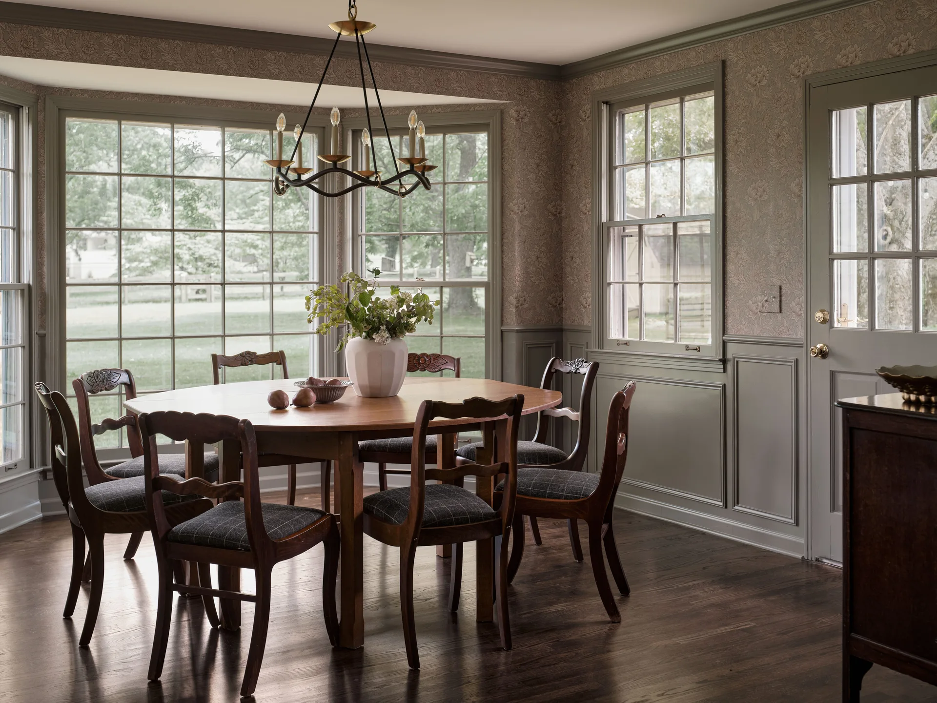 A traditional dining room features a round wooden table, matching chairs with plaid seats, olive green wainscoting, and patterned wallpaper.