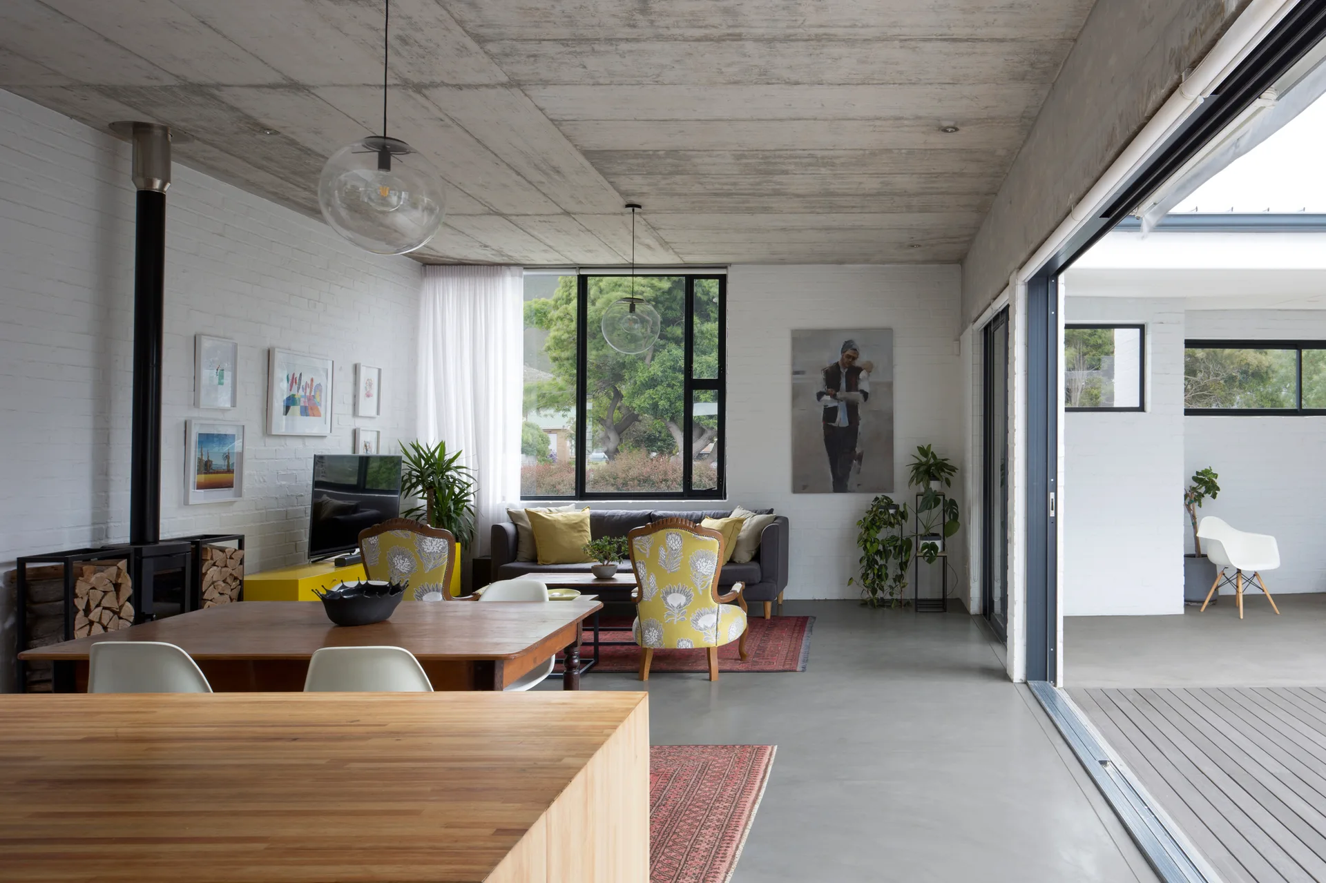 An open-plan living and dining space features a grey sofa, yellow armchairs, a wooden dining table, and a concrete ceiling.