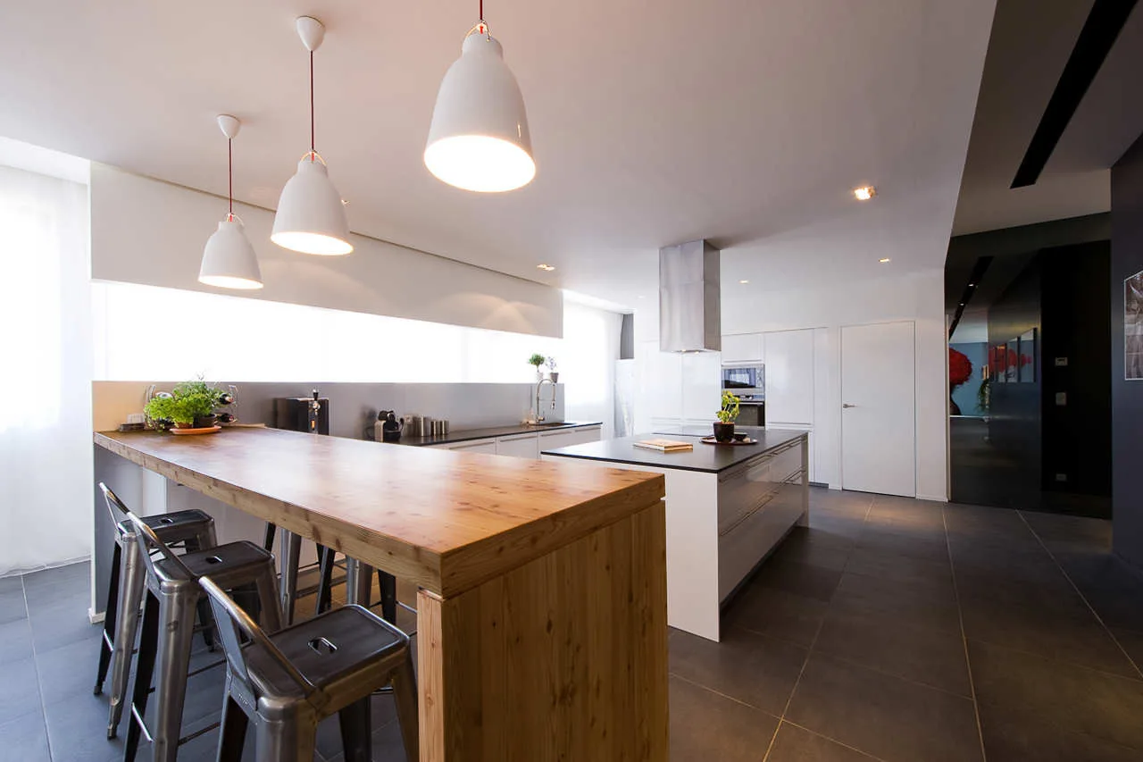 A modern kitchen features a long natural wood breakfast bar with industrial metal stools and white pendant lights hanging above.