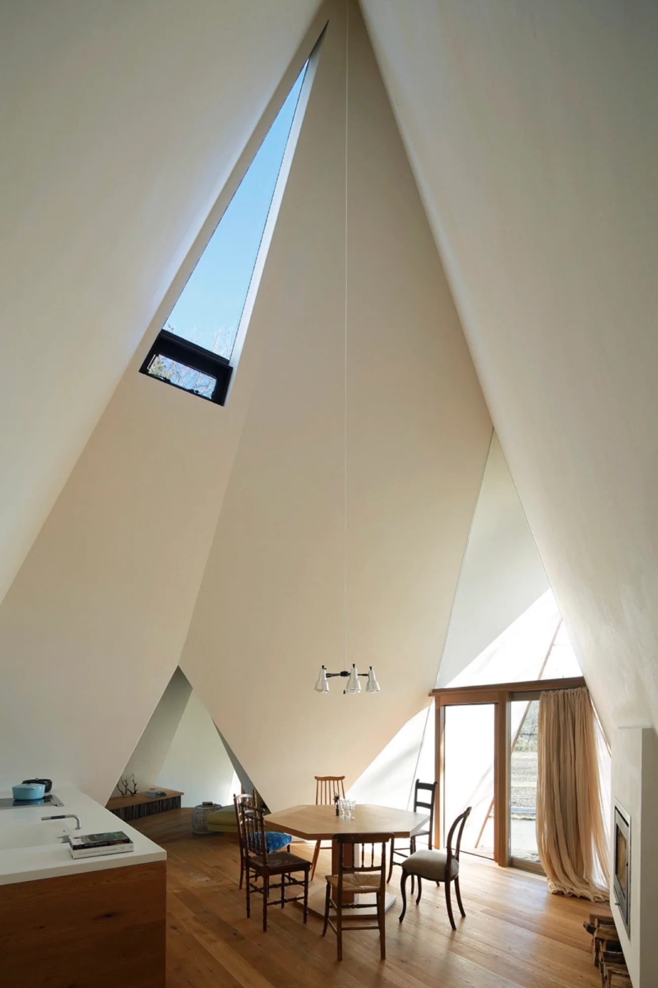 A minimalist dining room features a hexagonal wooden table and mismatched chairs set under a high, angular white vaulted ceiling with a skylight.