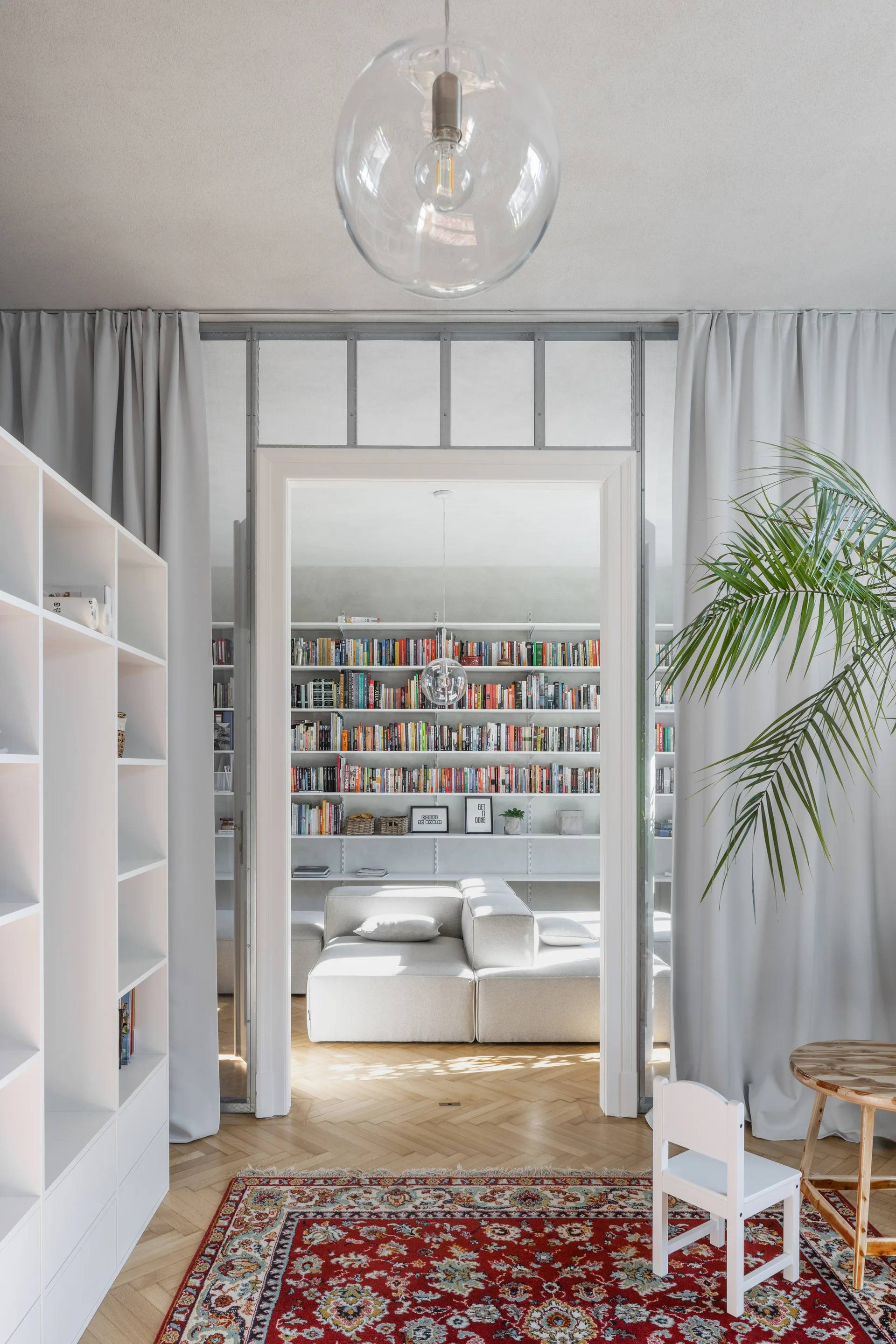 A view through a glass partition doorway into an adjacent room with a modular sofa and floor-to-ceiling white bookshelves over herringbone floors.