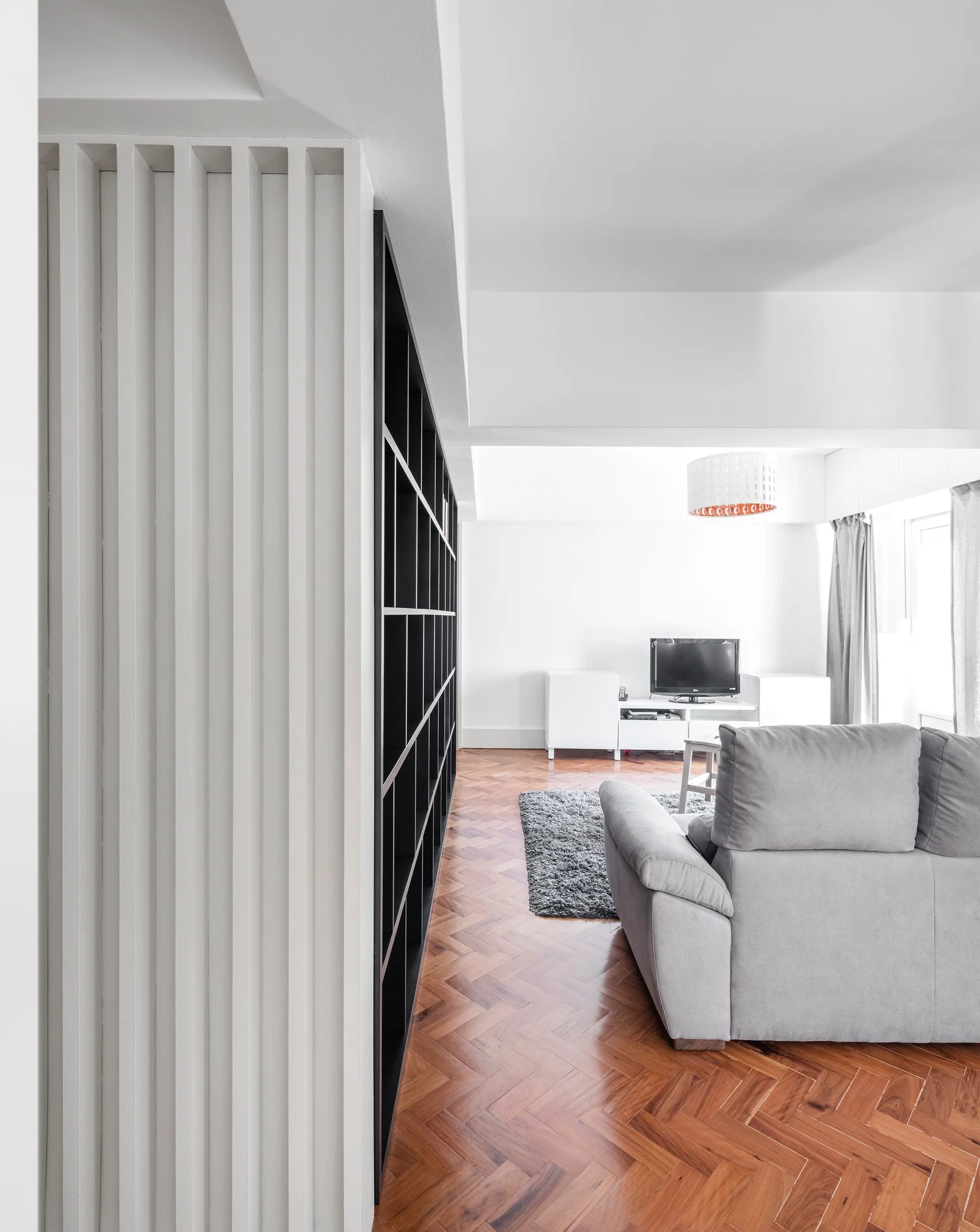 A modern living room features a grey fabric sofa on herringbone wood flooring next to a large black grid bookshelf and a white slatted wall.