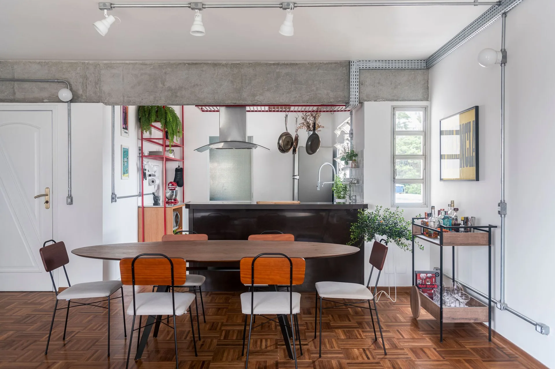 An industrial dining area features an oval wood table with chairs on parquet flooring, next to an open kitchen with an exposed concrete beam.