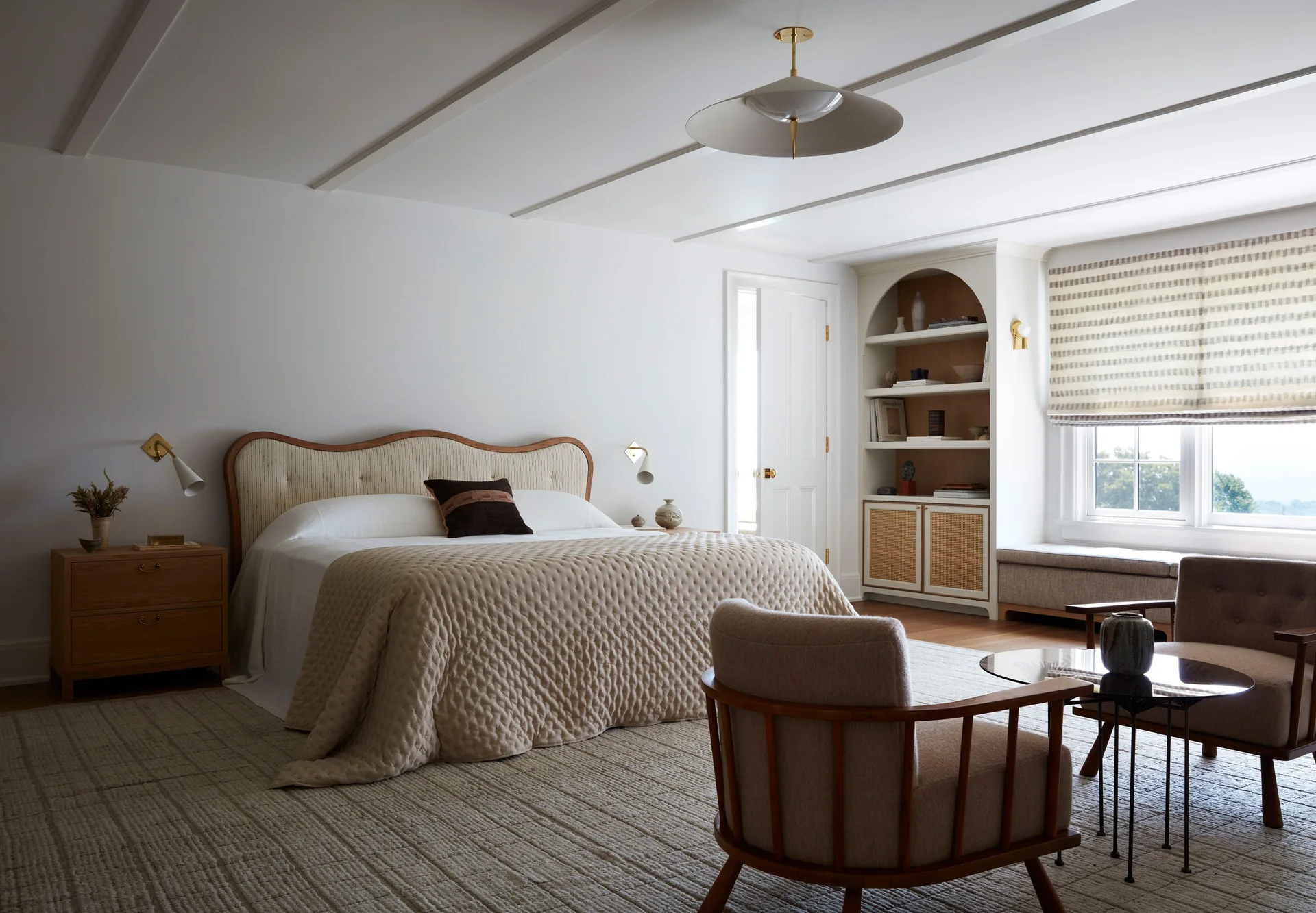 A neutral-toned bedroom featuring an upholstered bed, a large arched built-in shelving unit, wooden armchairs, and a striped window shade.