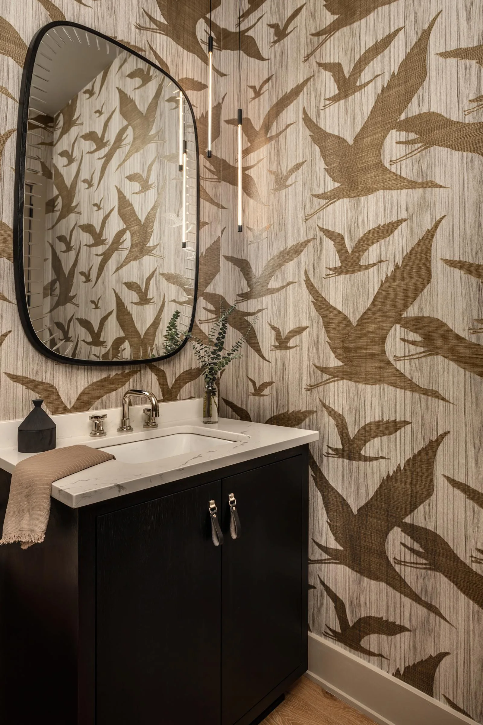 A modern powder room features a dark vanity with a marble countertop, an oval mirror, and gold bird-patterned wallpaper.