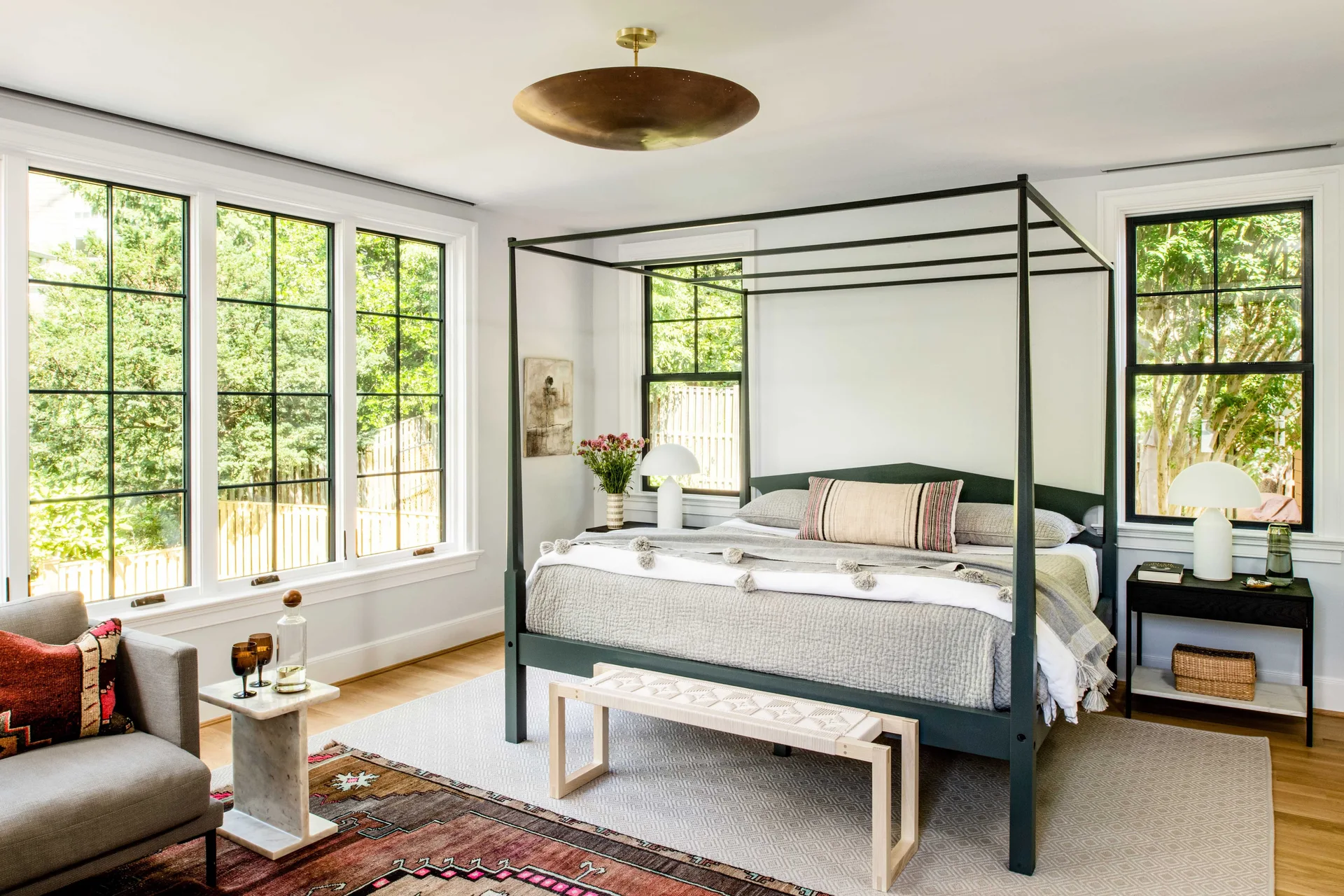 A modern bedroom featuring a black metal canopy bed, large black-frame windows, and a woven bench at the foot of the bed.