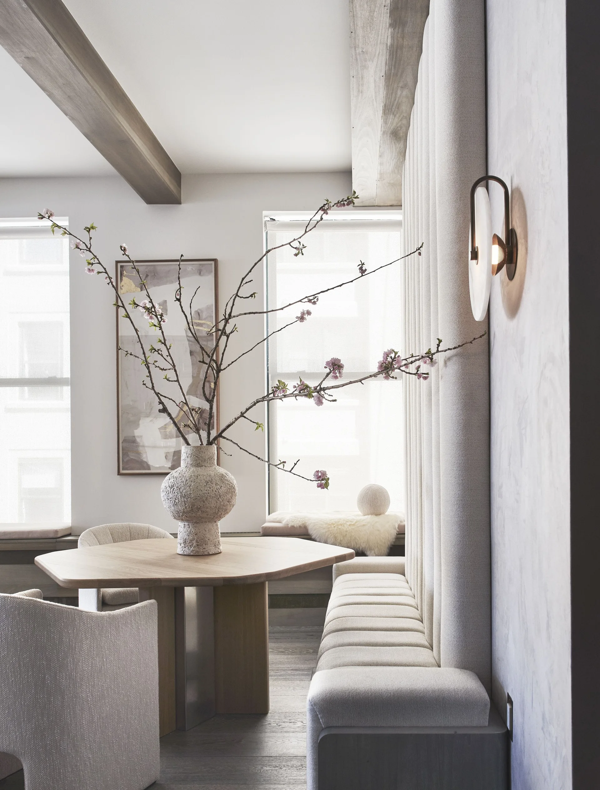 A modern dining nook featuring a wood table, a tall channel-tufted banquette, exposed ceiling beams, and a stone vase with flowering branches.