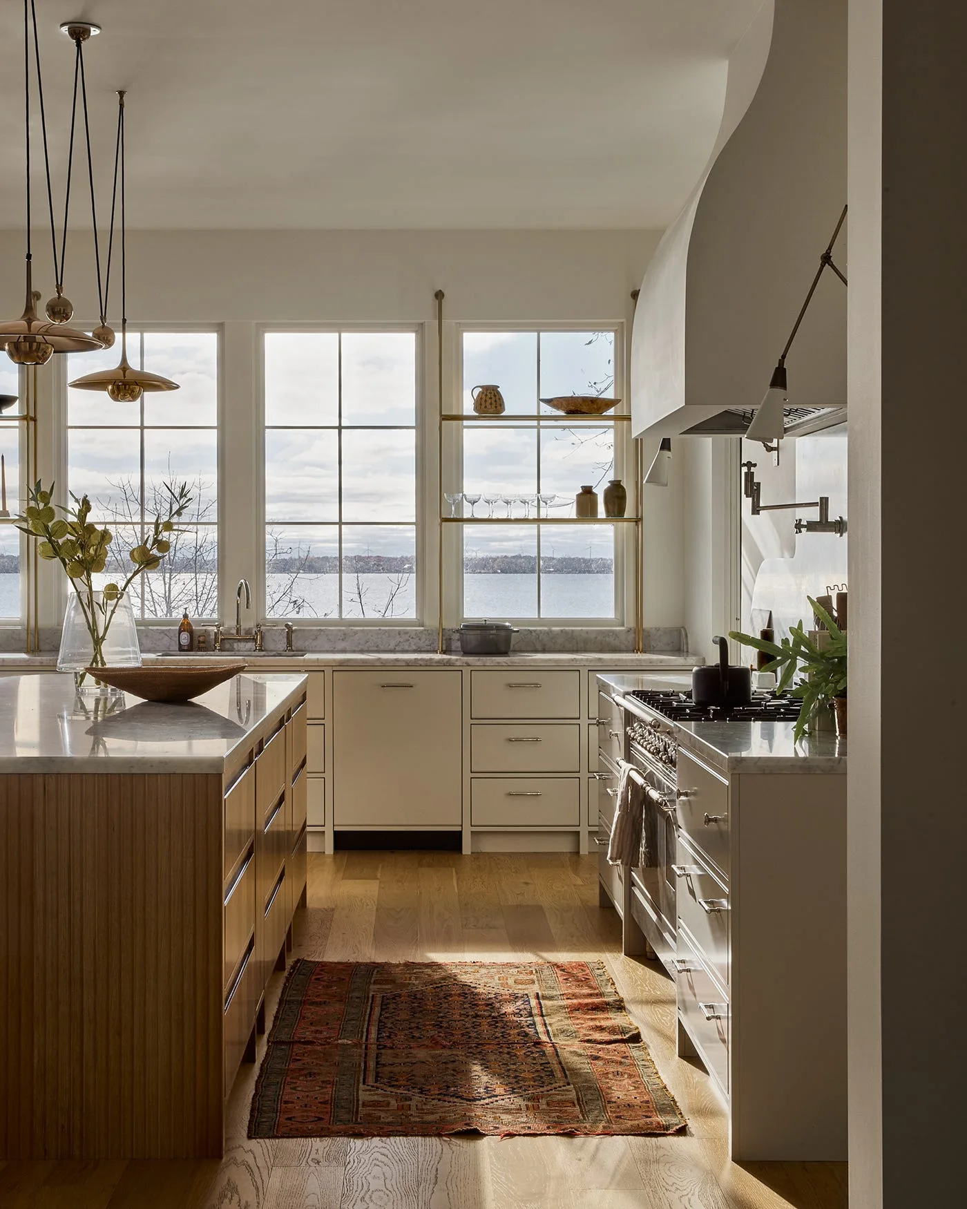 A bright kitchen features a wood-toned island with a marble top, large windows with brass shelves, and a range with a plaster hood.