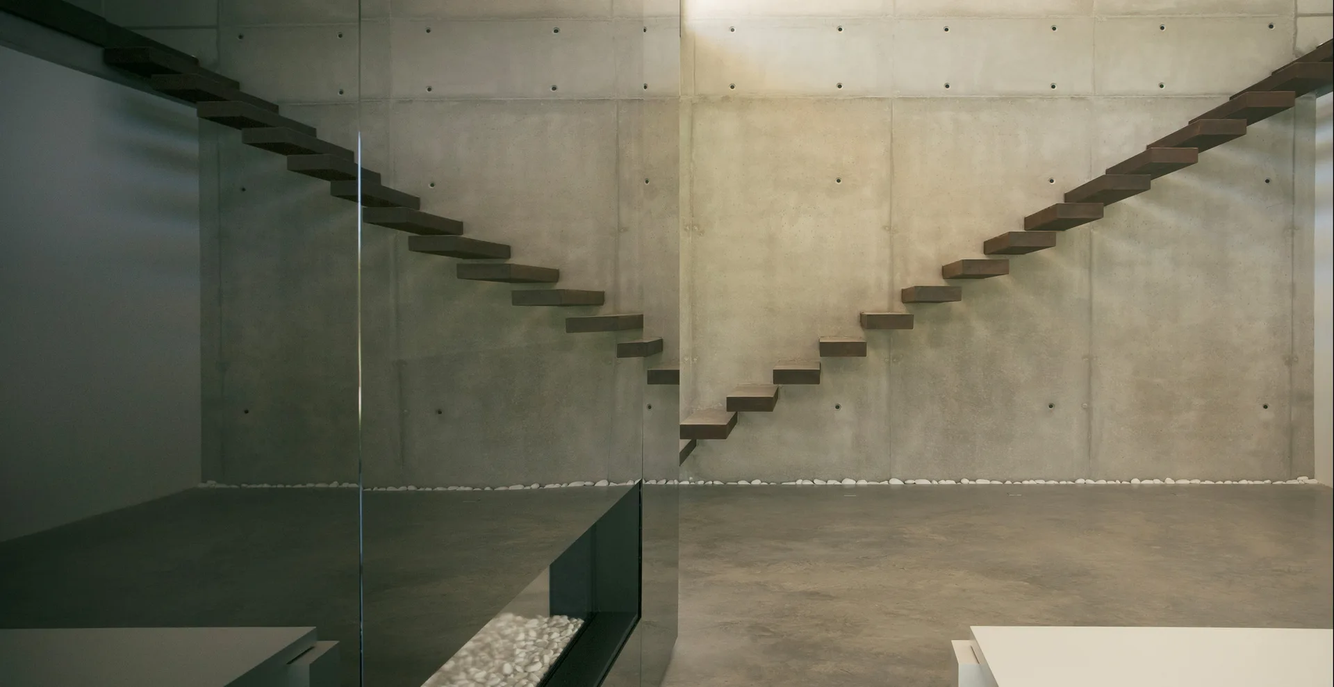 A minimalist interior featuring cantilevered wooden stairs set against an exposed concrete wall with a reflective glass partition and floor pebbles.