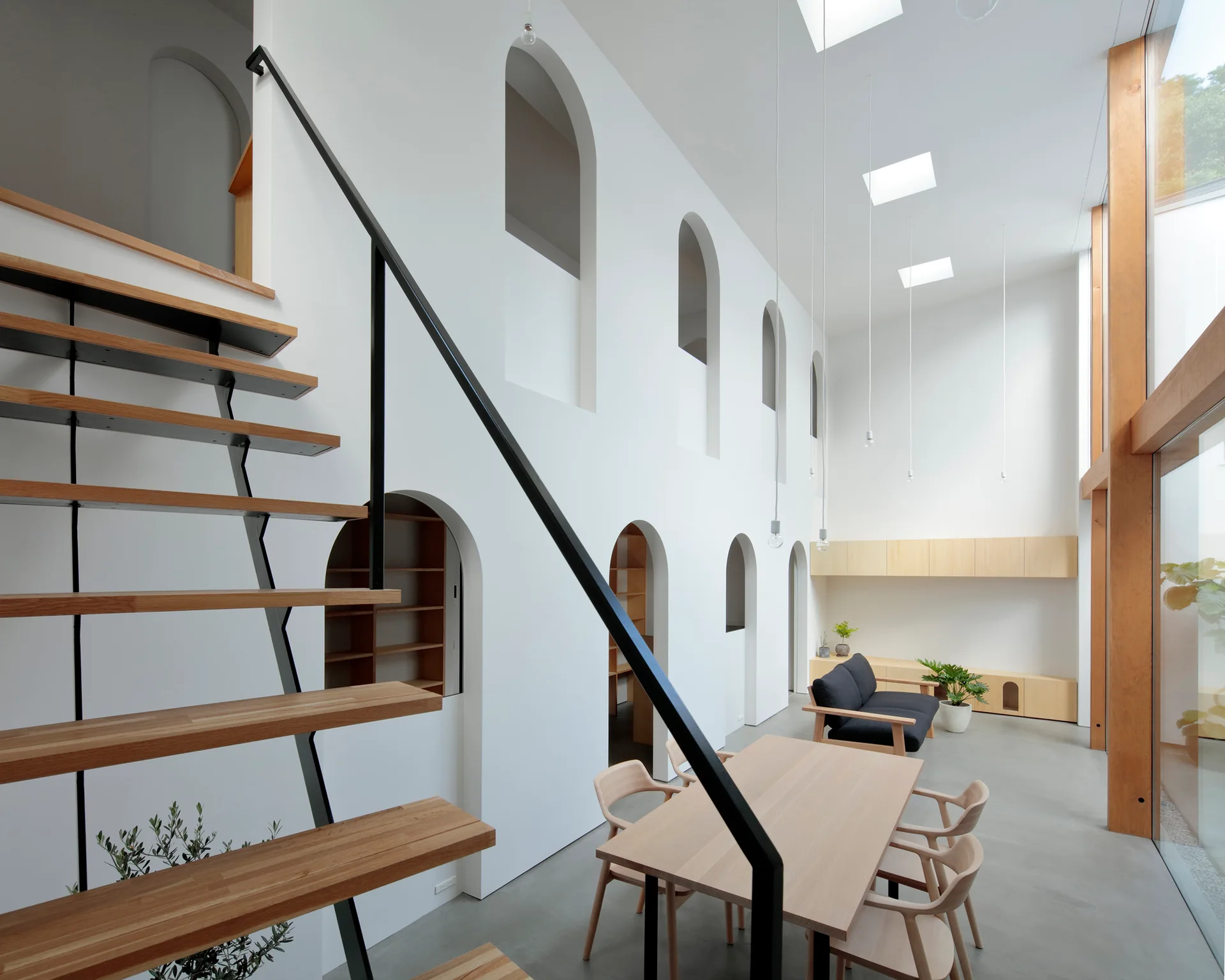 A minimalist double-height living room featuring a white wall with arched openings, a floating wood staircase, and a matching dining table set.