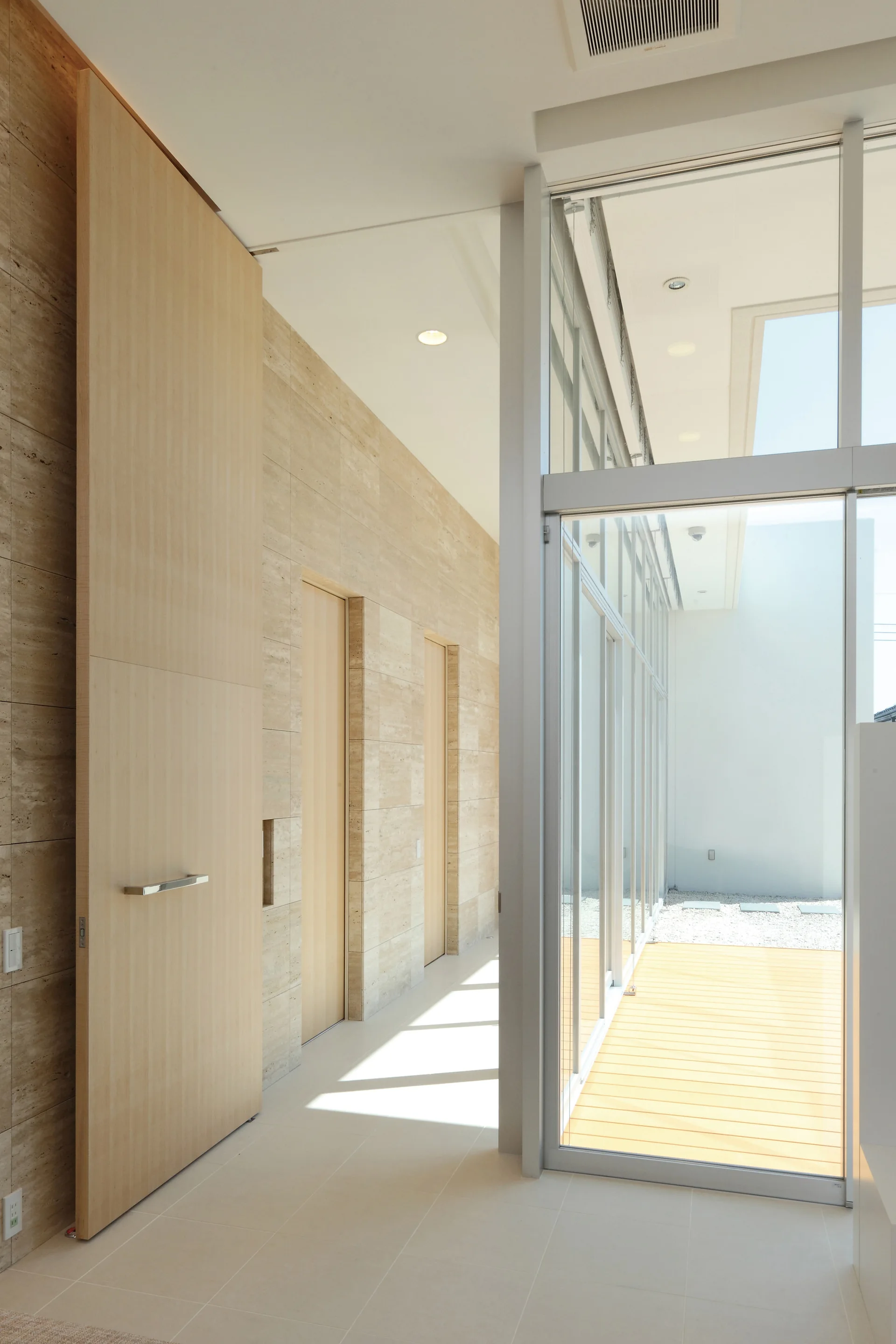 A bright minimalist hallway features travertine stone walls with inset wooden doors next to a floor-to-ceiling glass wall leading to a deck.