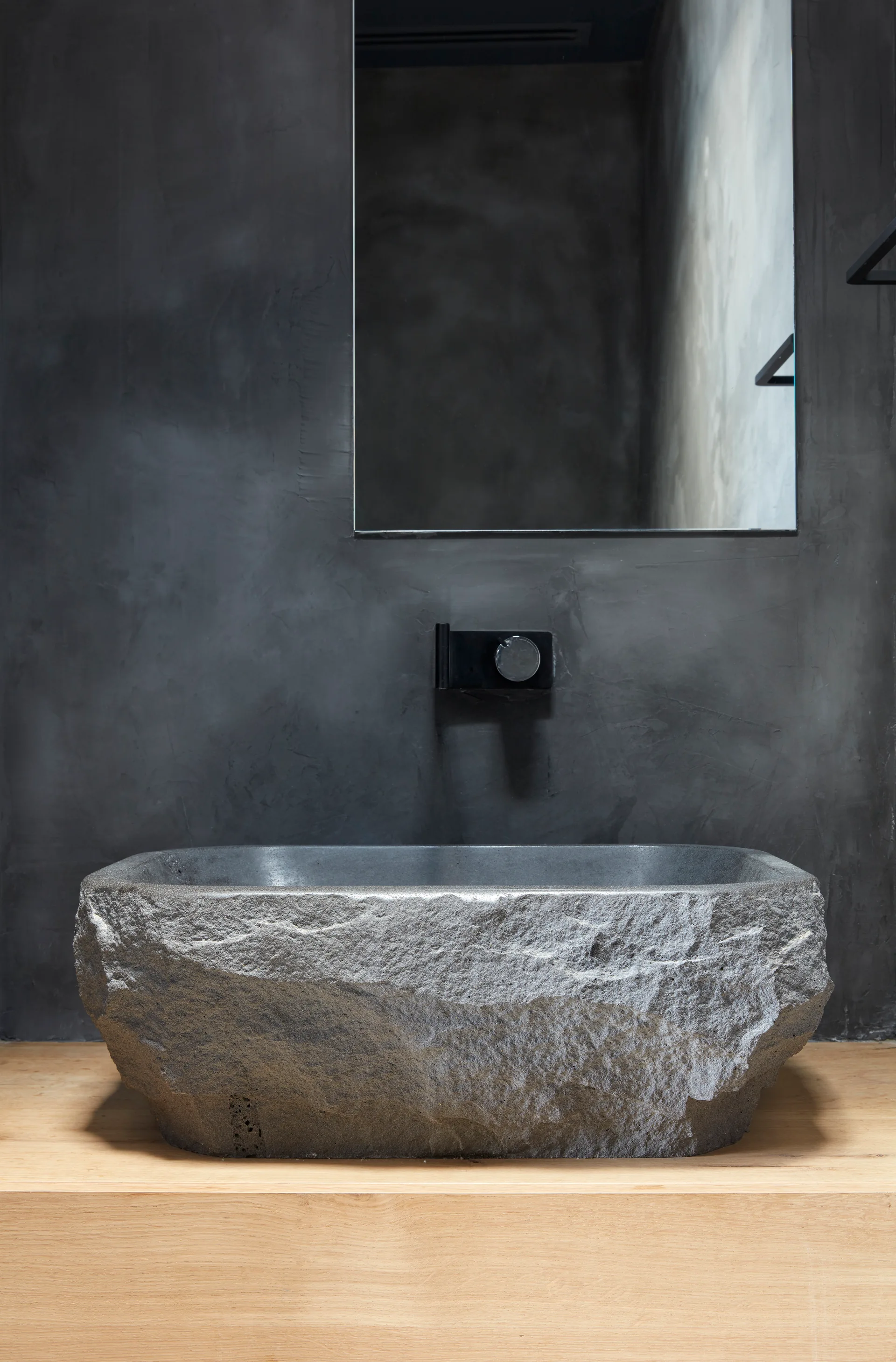 A rough-textured gray stone basin sits on a light wood countertop against a dark concrete wall with a wall-mounted black faucet.