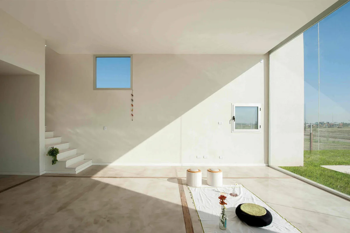 A minimalist off-white living room featuring a large floor-to-ceiling glass wall, white staircase, polished concrete floors, and low floor seating.