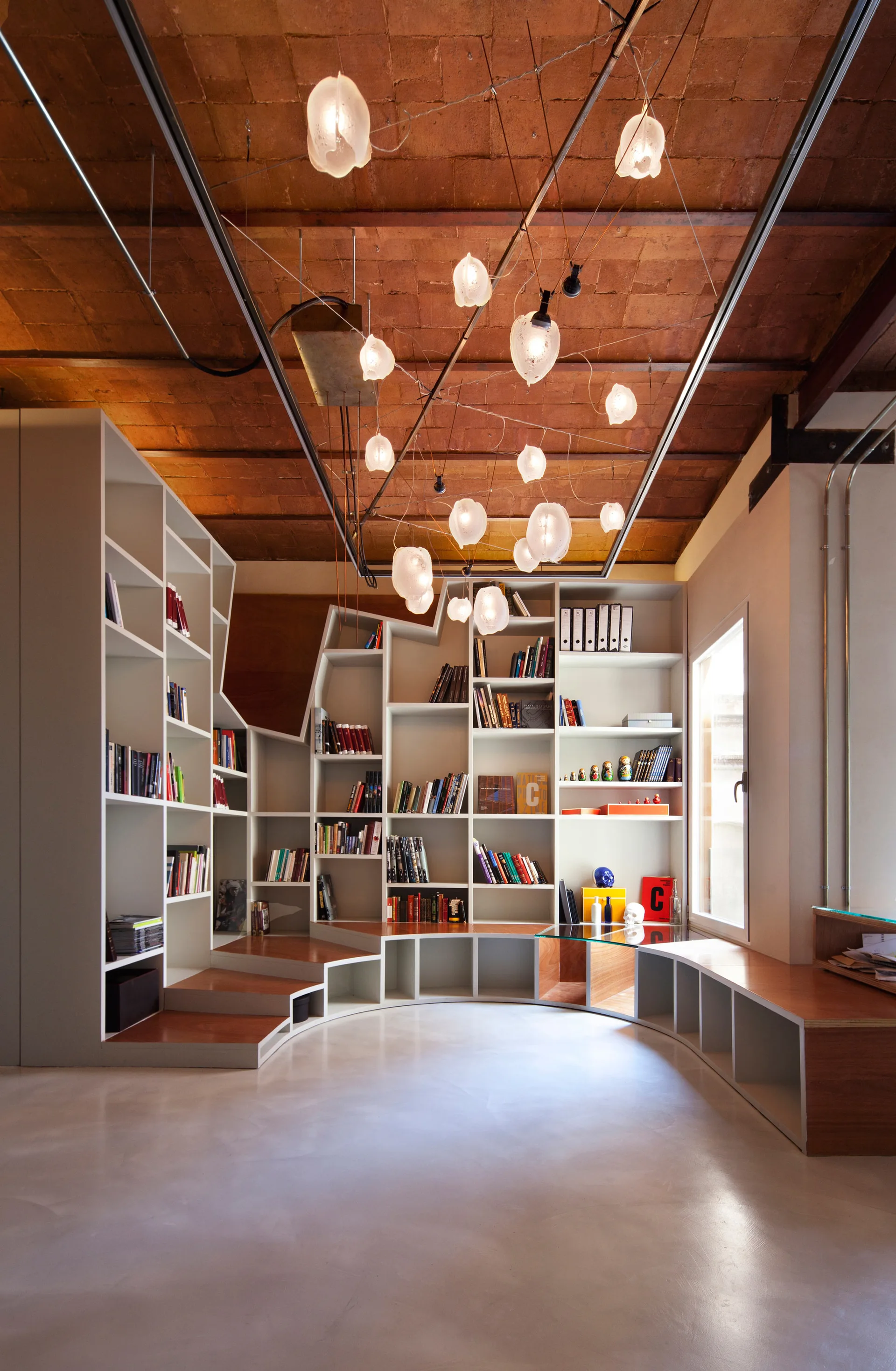 A modern library room features a custom curved white and wood bookshelf beneath a vaulted red brick ceiling and a cluster of glowing orb pendant lights.