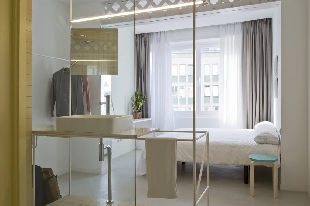 A modern studio apartment featuring an integrated open white metal vanity with a wash sink next to a bed with white linen.