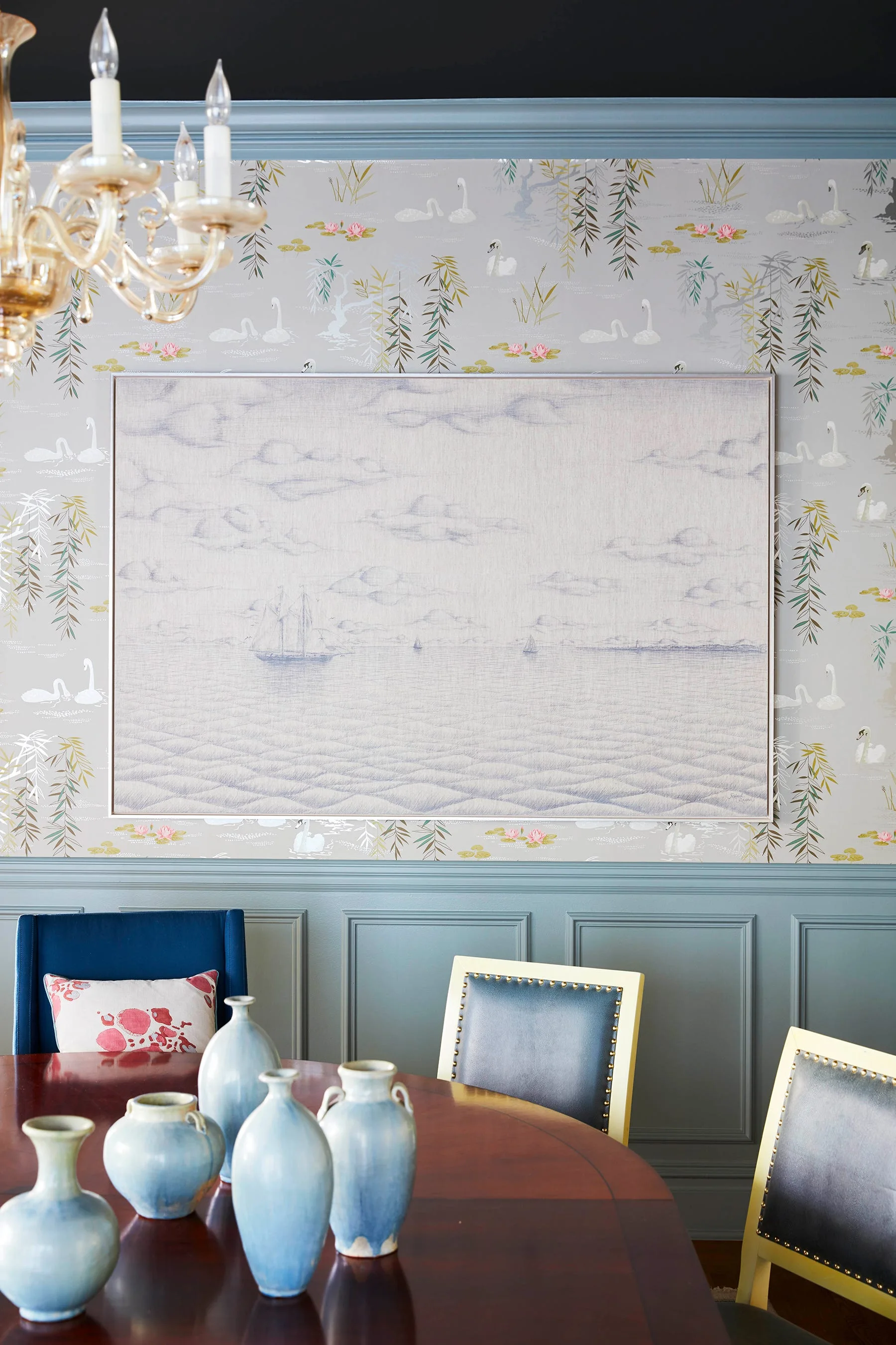 A dining room featuring a wooden table with blue ceramic vases, paneled wainscoting, and swan-motif wallpaper behind a large seascape drawing.