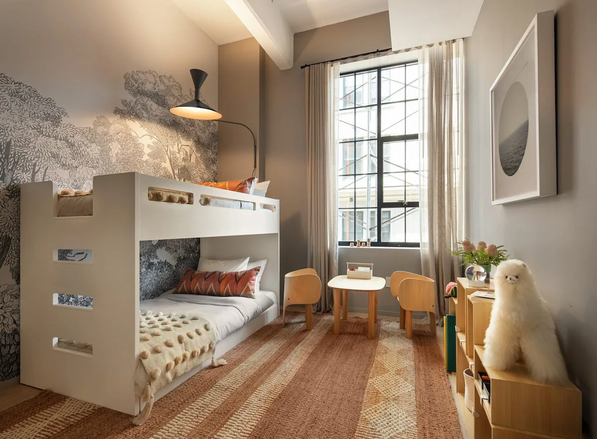 A children's bedroom features a white bunk bed against a black and white forest mural wall next to a wooden play table.