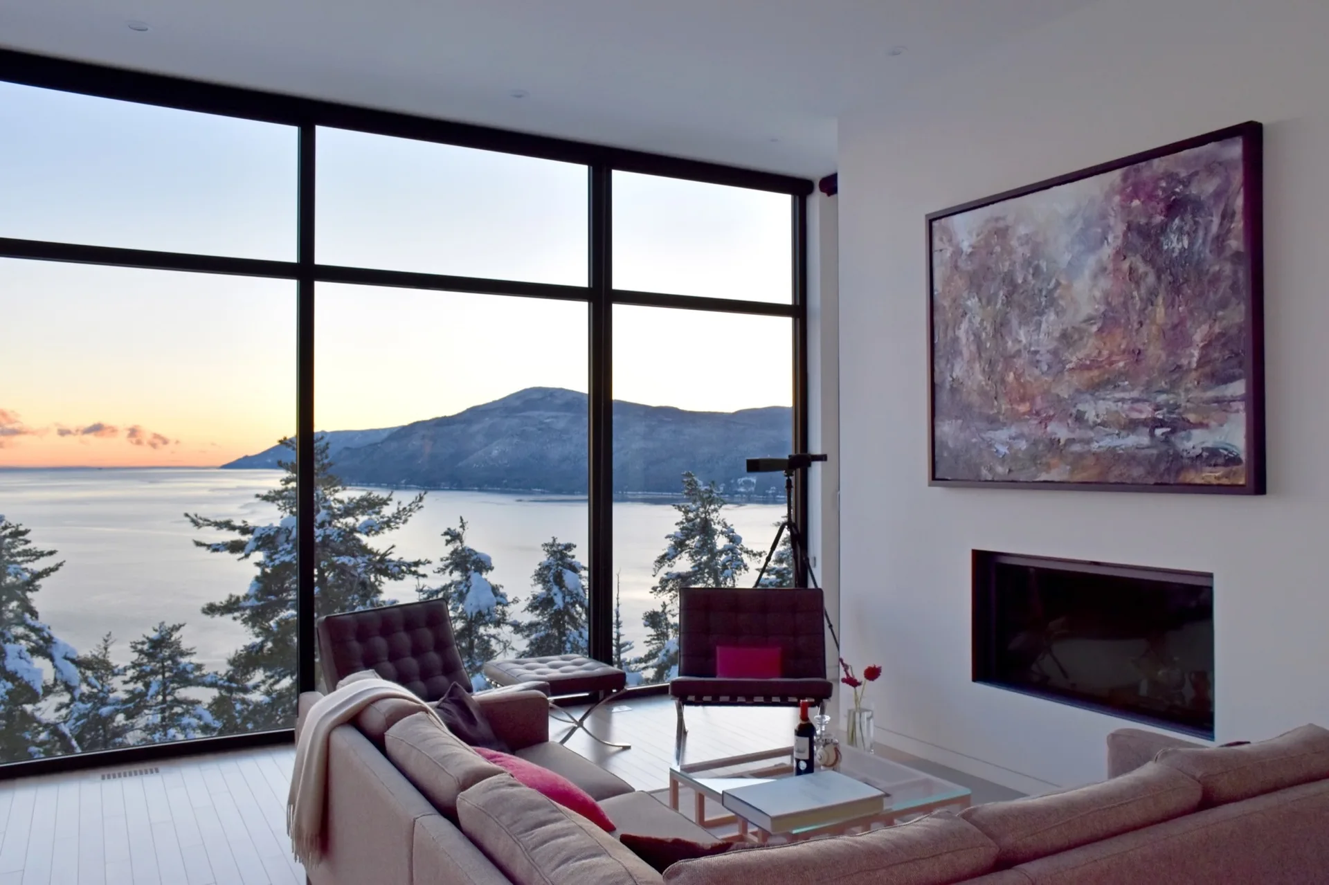 A modern living room features a beige sectional sofa, a recessed fireplace, and a large black-framed window overlooking a snowy lakeside mountain view.