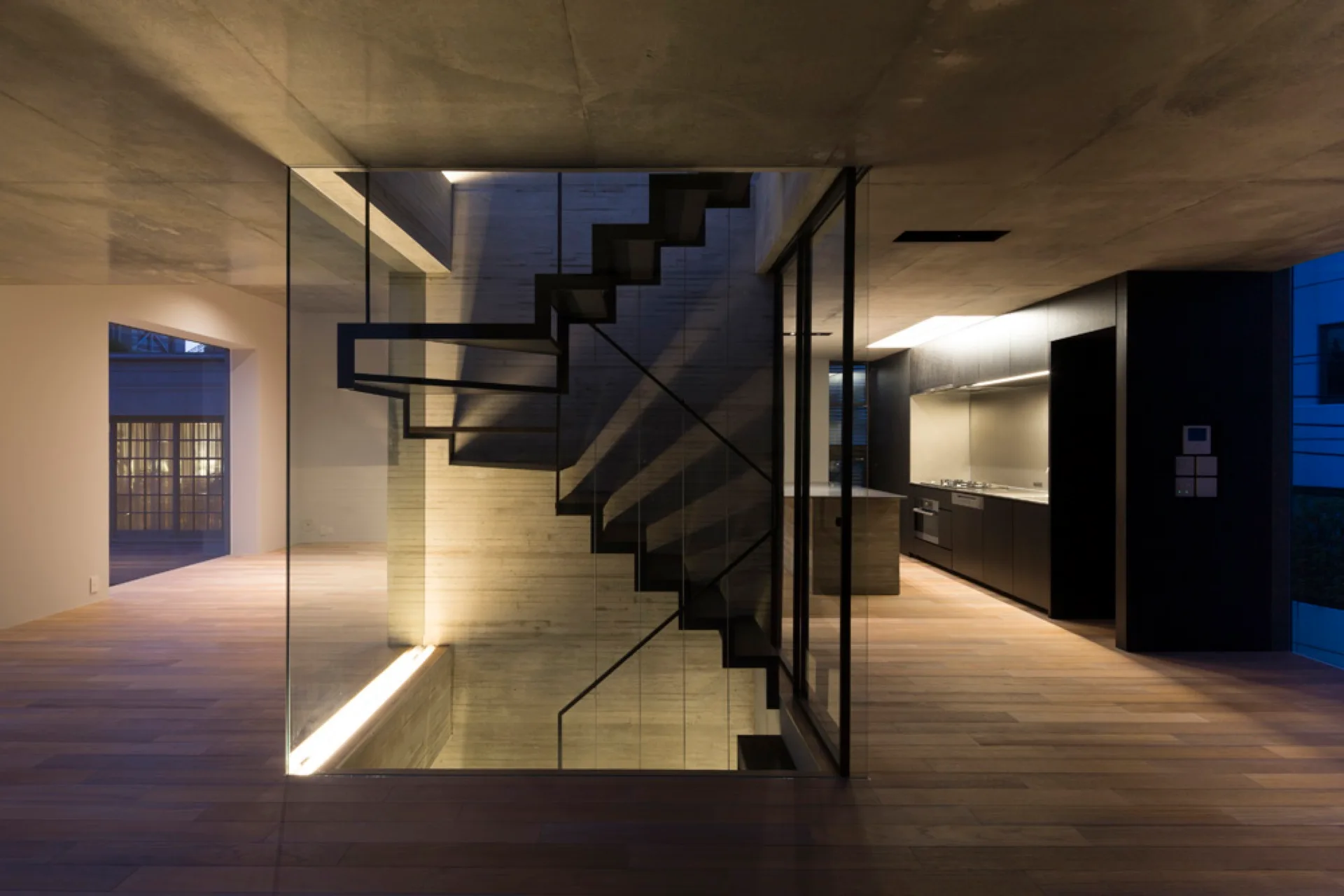 A minimalist black floating staircase surrounded by glass panels stands on wood floors against a raw concrete wall.