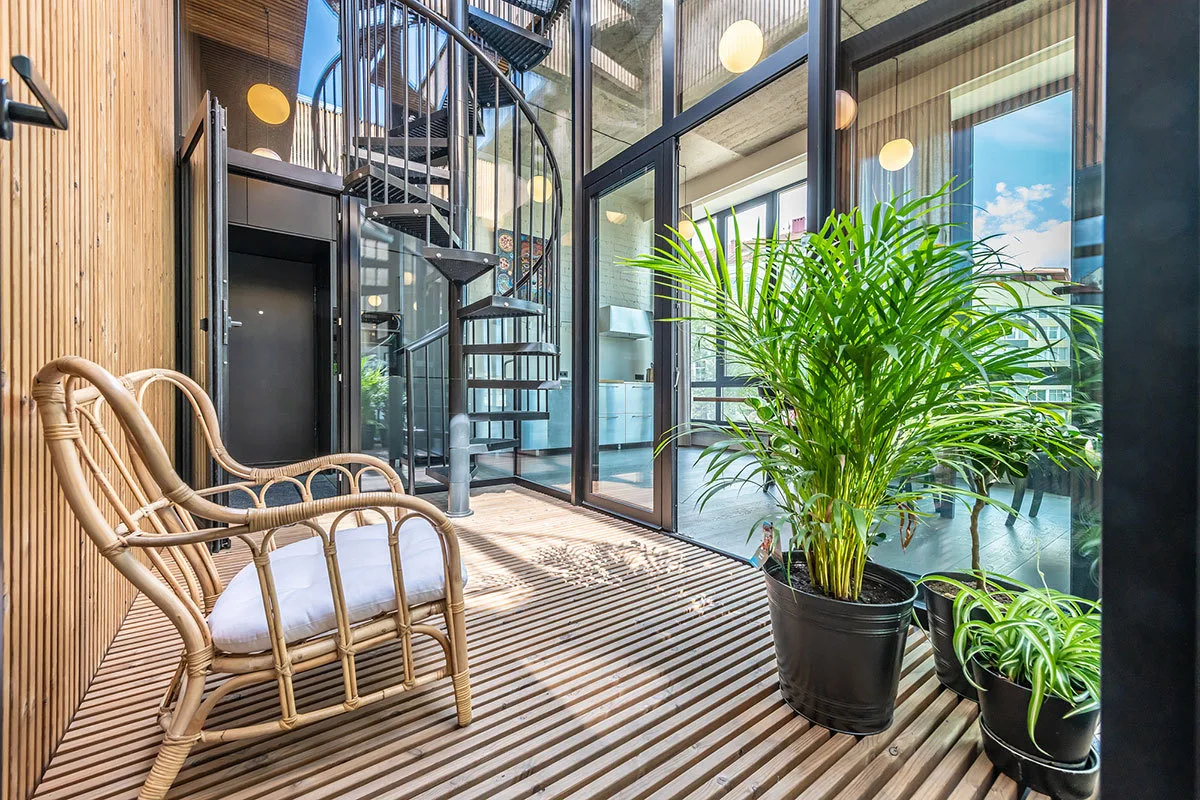 A modern sunroom features a black metal spiral staircase, wood-slatted floors and walls, a rattan armchair, and tall potted plants.