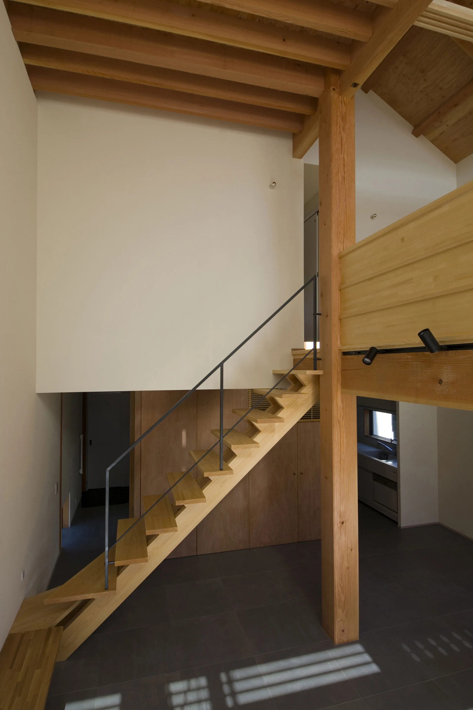 A modern wooden staircase with a slim metal railing ascends next to a tall timber column in a home with exposed ceiling beams and dark tile flooring.