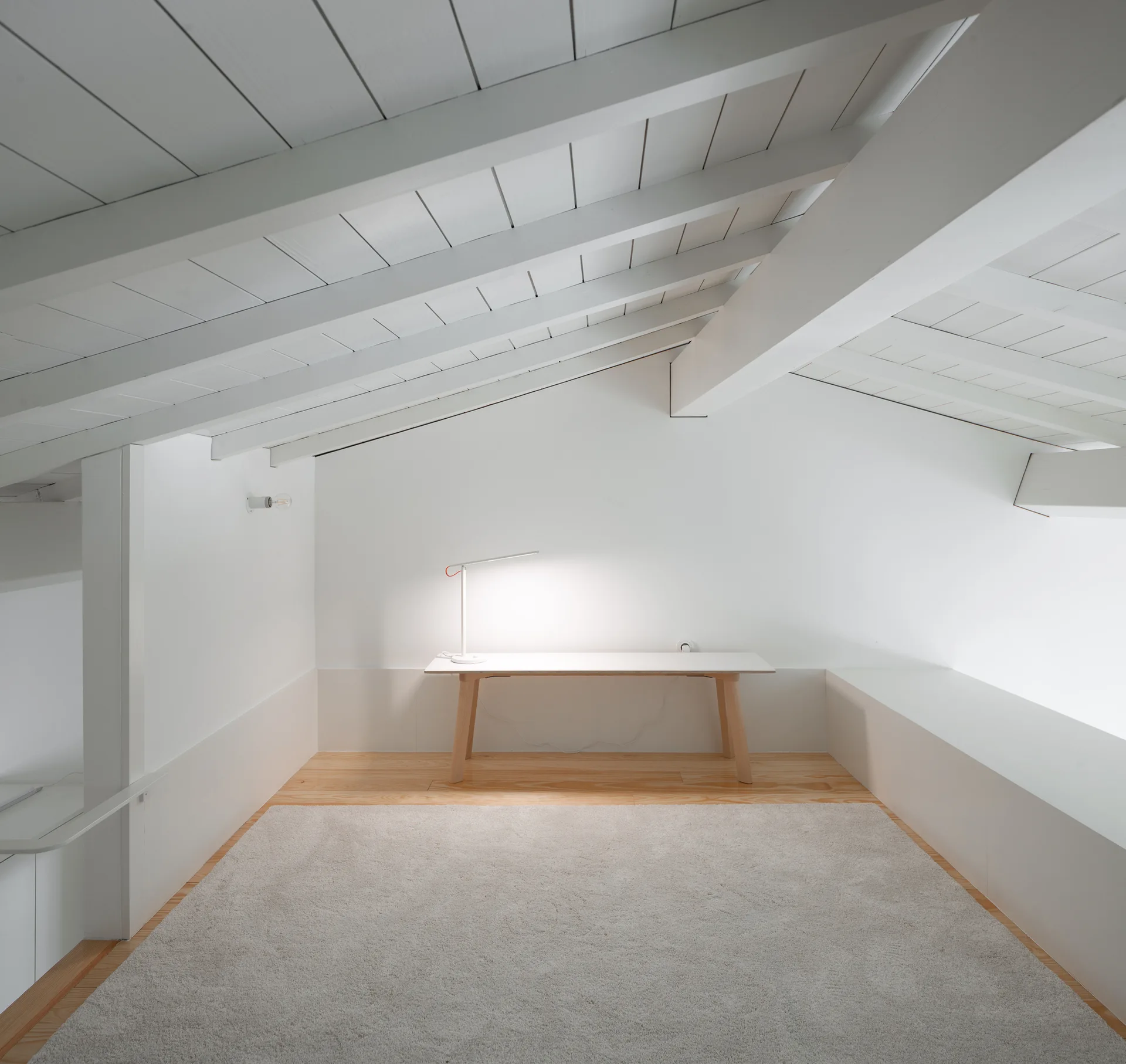A minimalist attic home office features a white desk, table lamp, gray rug, and a slanted ceiling with painted wooden beams.