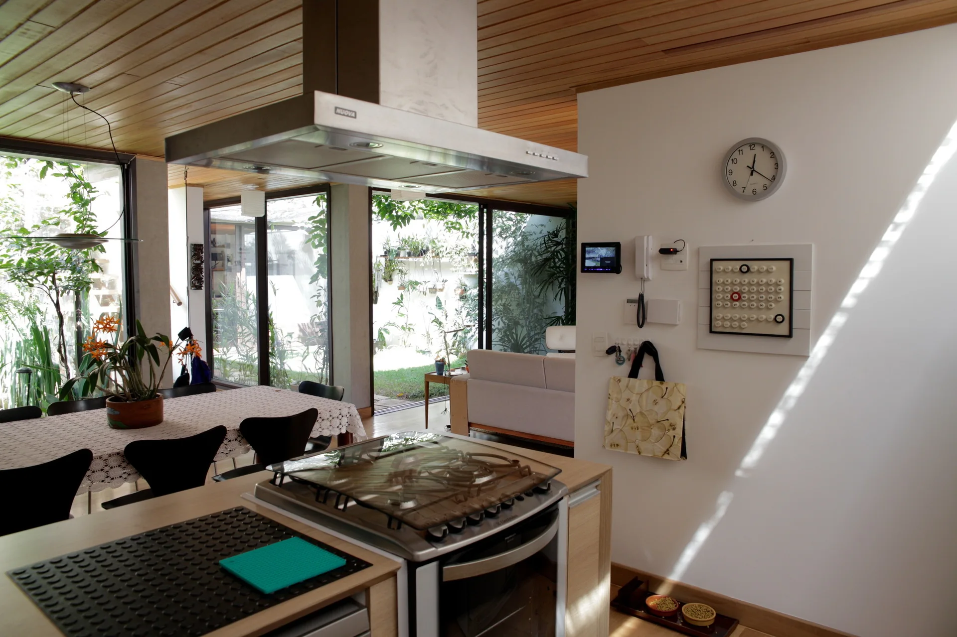 A modern open-plan kitchen featuring a stainless steel range hood, wooden plank ceiling, and glass doors opening to a lush garden.