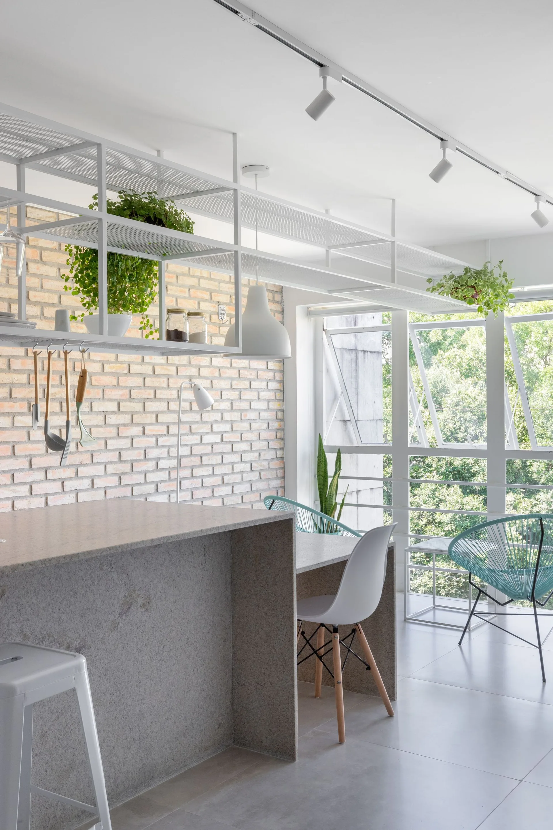 A modern kitchen featuring a textured stone island, an exposed brick wall, and a white ceiling-mounted metal mesh shelf with hanging plants.
