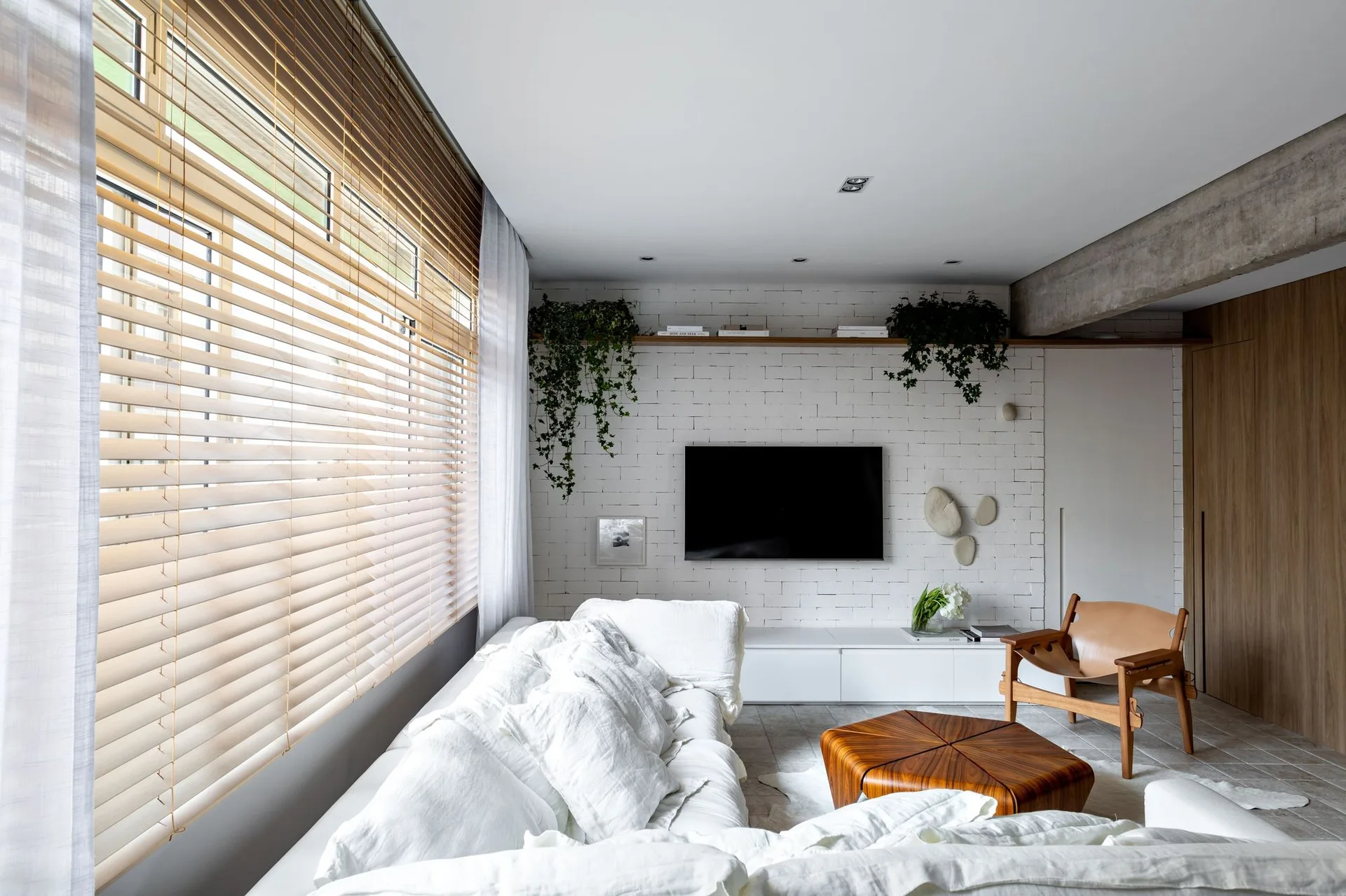 A modern living room features a white textured sofa, a sculptural wood coffee table, a white painted brick wall, and natural light from wood blinds.