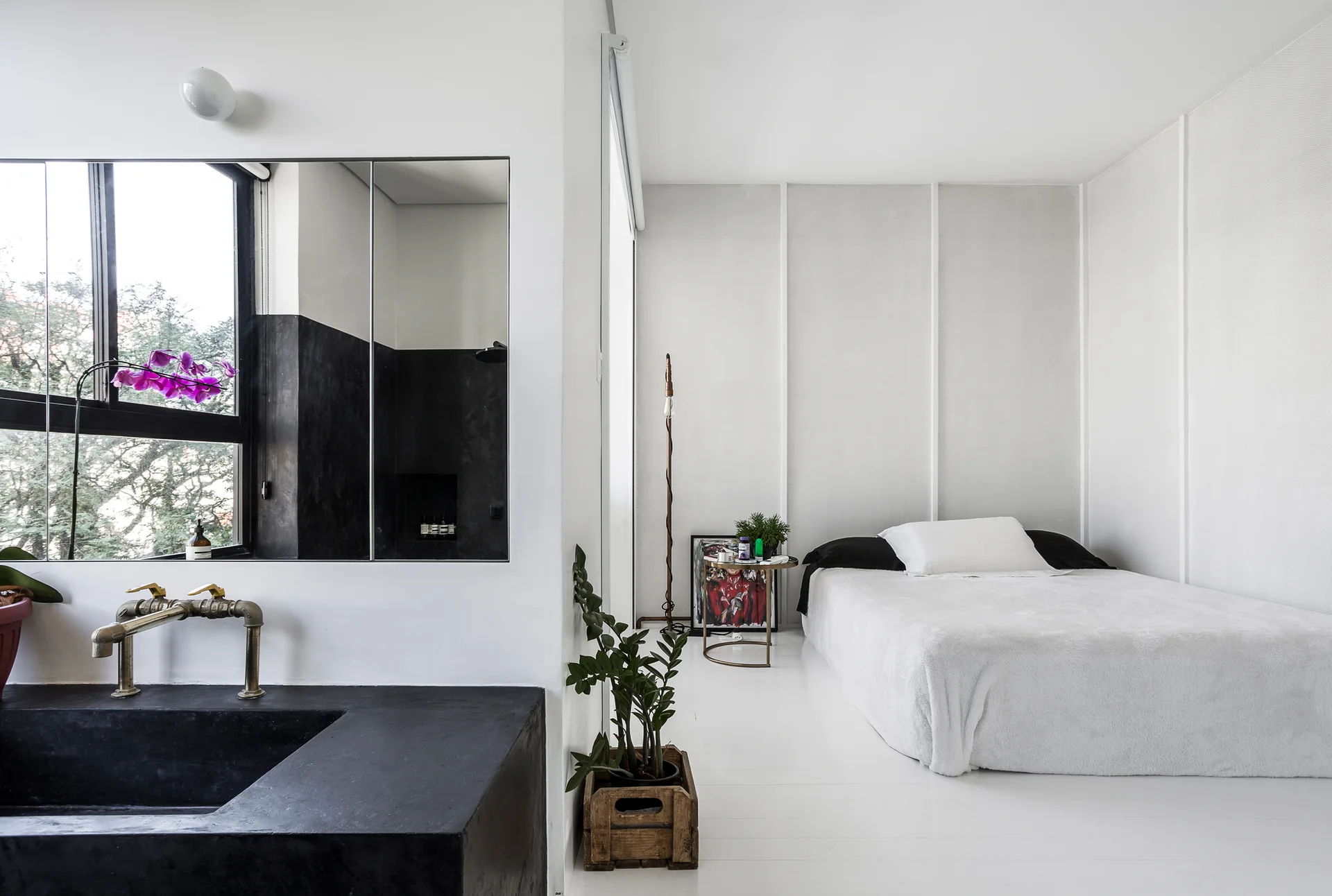 A minimalist bedroom with a low profile bed and white paneled walls adjacent to an open bathroom featuring a black stone vanity and brass faucet.