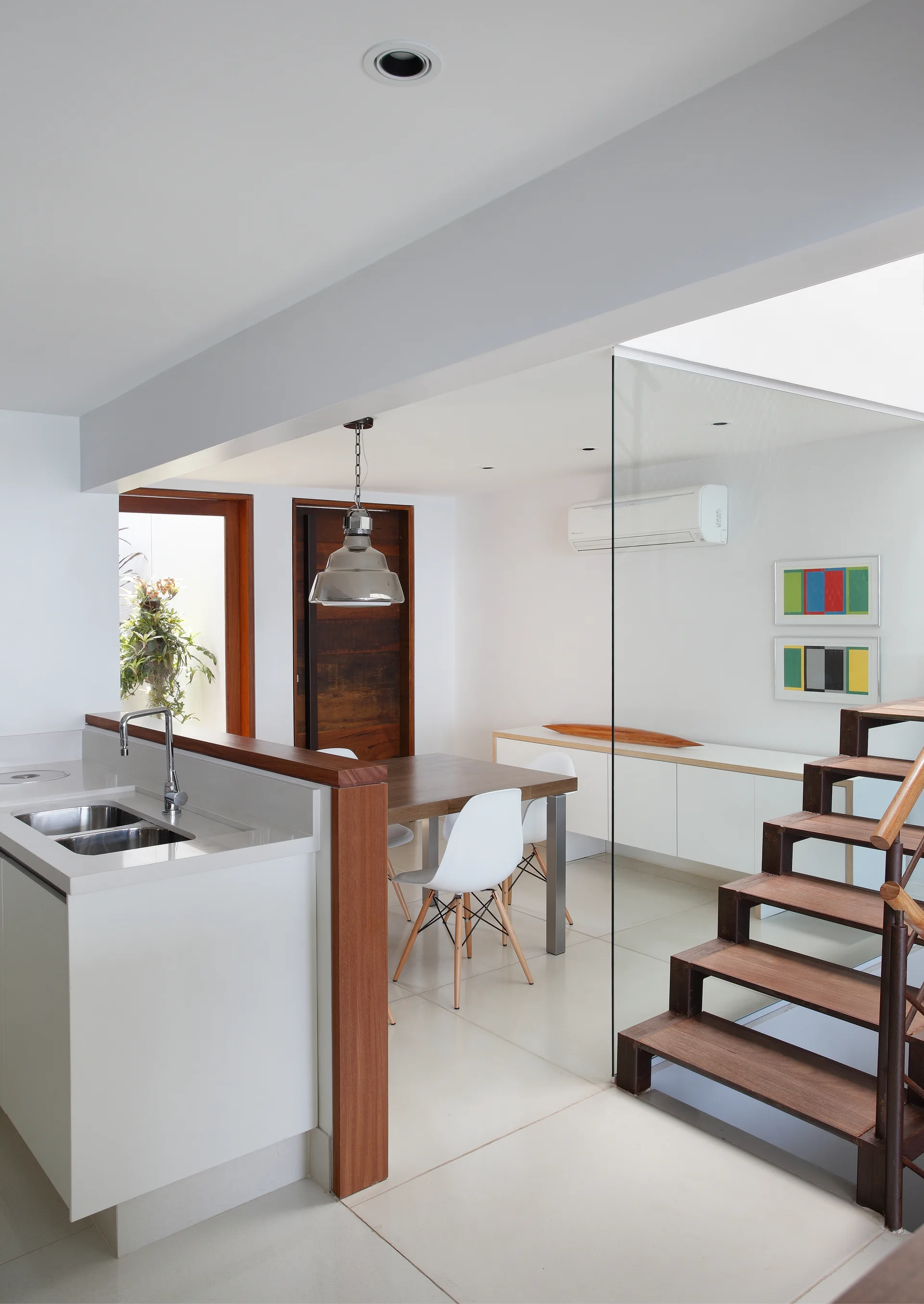 A modern open-plan dining room features a wooden table, white chairs, and an open-riser staircase separated by a frameless glass partition.