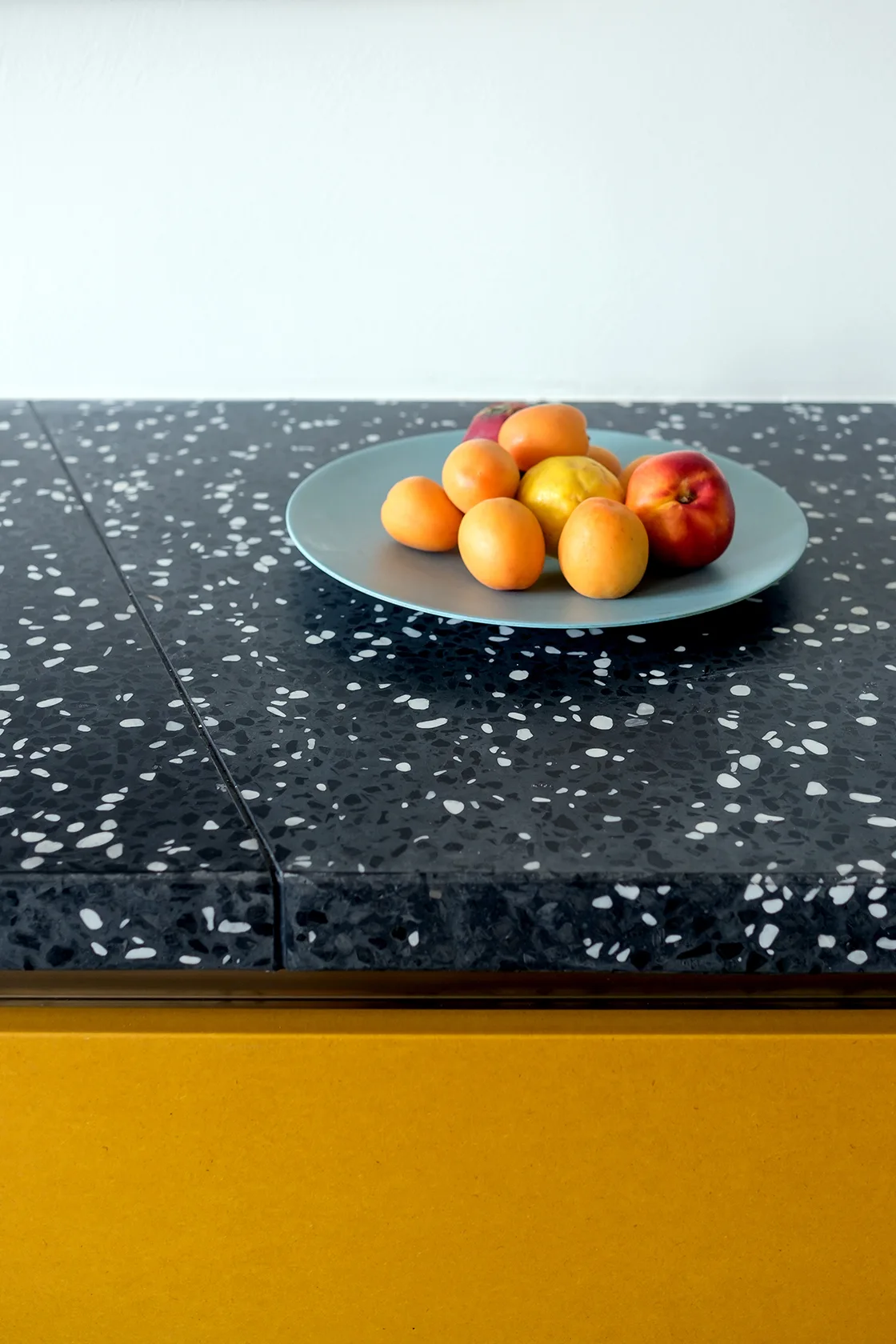 A speckled black terrazzo countertop sits above a solid matte yellow cabinet panel, with a light blue fruit plate resting on top.
