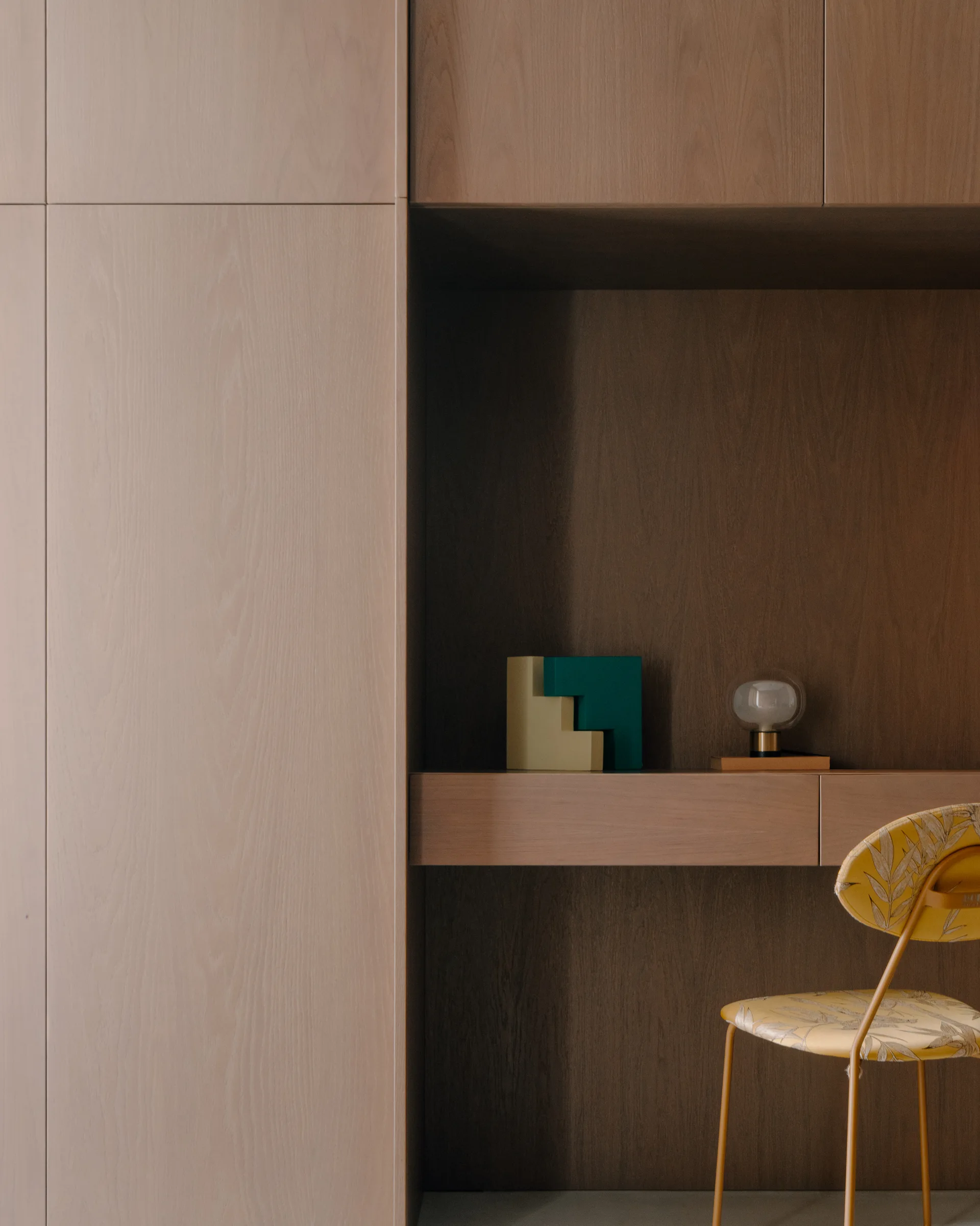 A minimalist home office desk recessed into large wood cabinetry, paired with a yellow leaf-patterned chair and geometric desk decor.